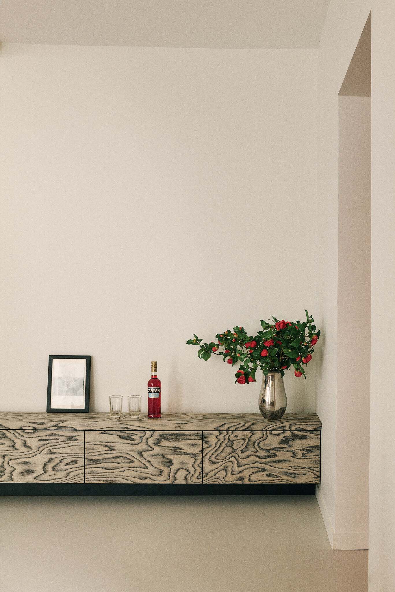 Floating burl-grained plywood sideboard holding a framed print, a red bottle, two glasses and camellia branches in a silver vase