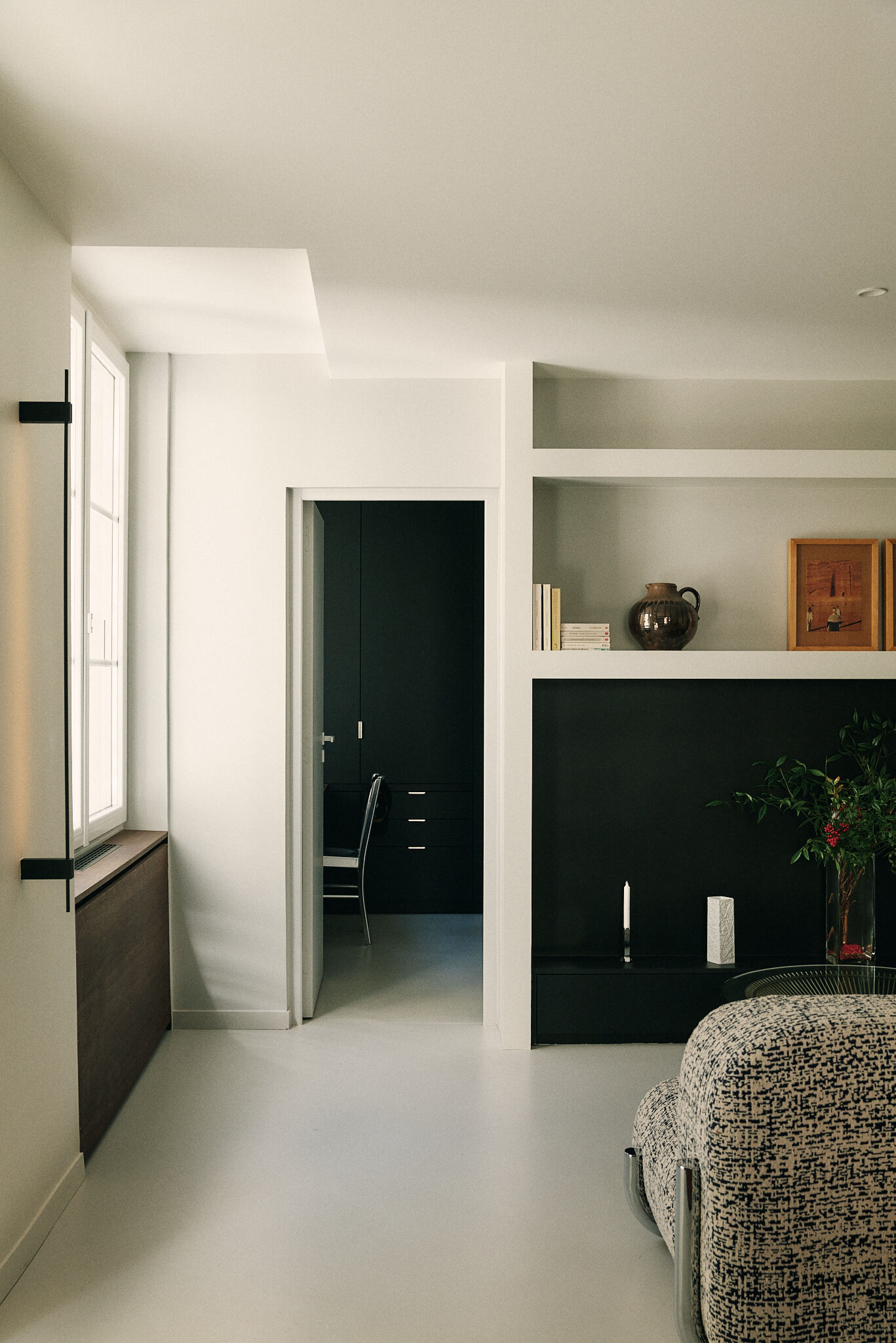 Living room corner with a black built-in shelf unit, an open doorway to a dark study and soft window light