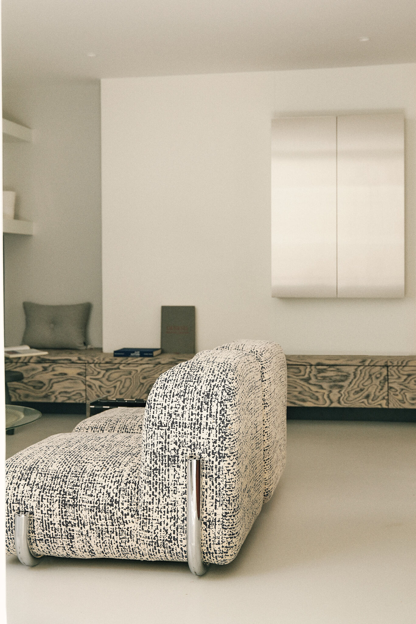 Back of a boucle-upholstered lounge chair with chrome legs, facing a long wood-grain sideboard and a steel wall cabinet