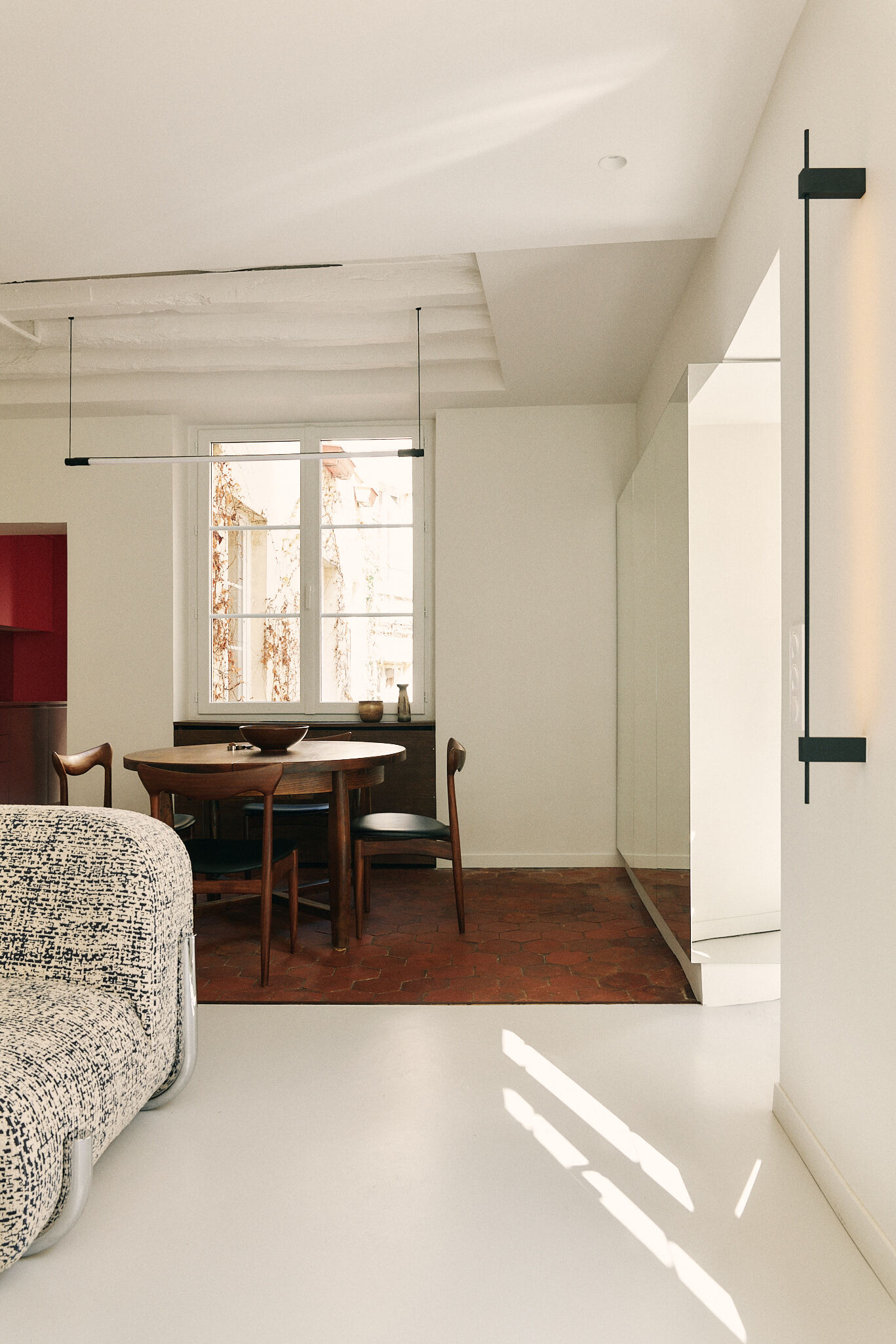 Dining area with a round timber table and teak chairs on terracotta tiles, lit by sunlight from a tall window