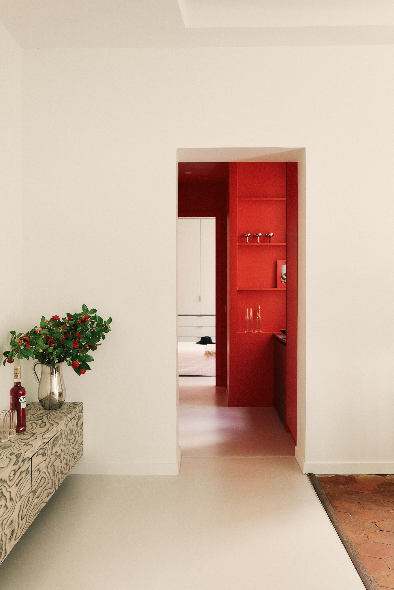 Doorway framing a saturated red shelving niche beyond the sideboard, with camellia branches in a vase and terracotta tile edge