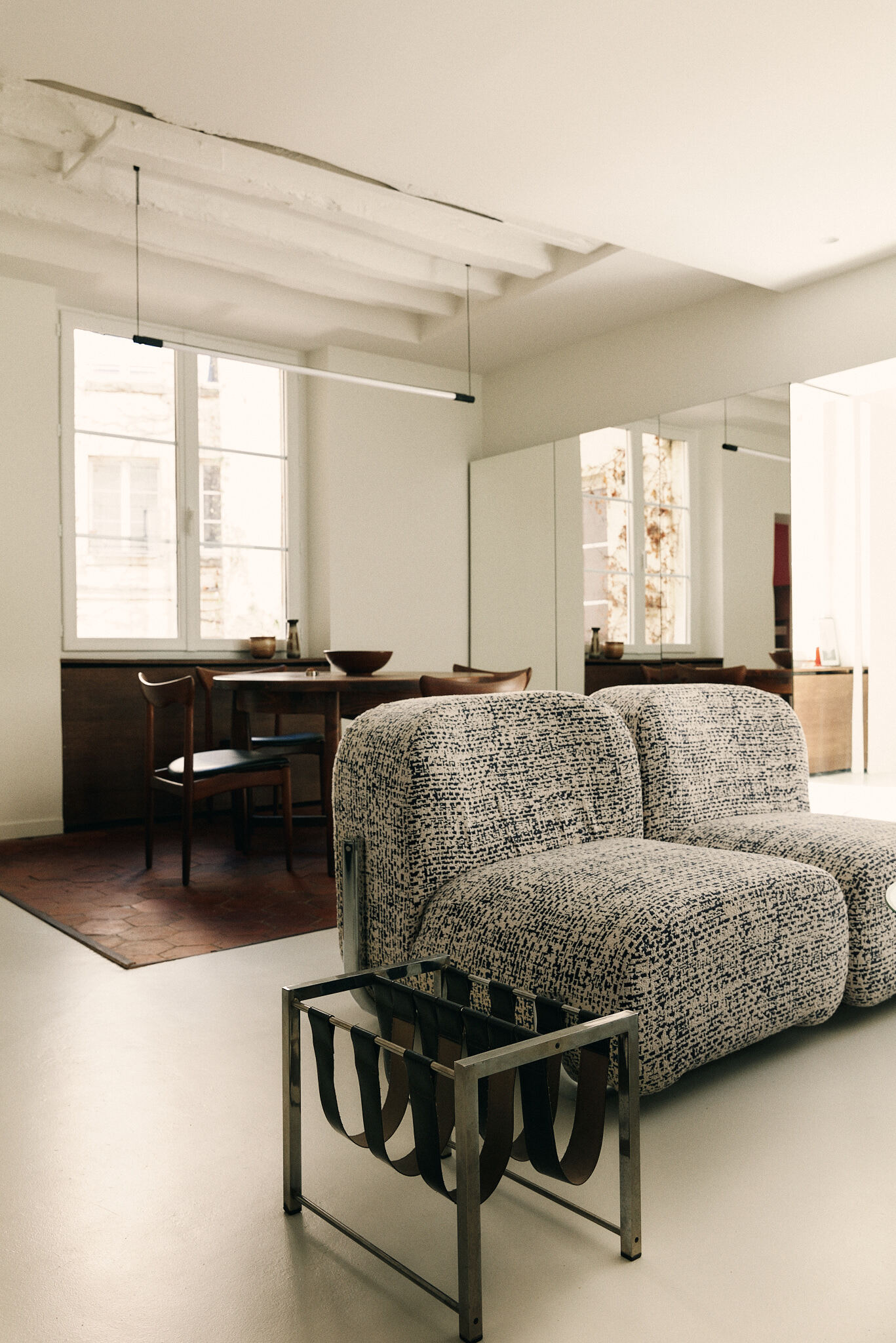 Boucle lounge seating and a leather-strap steel magazine rack in front of a dining table, with daylight from a tall window