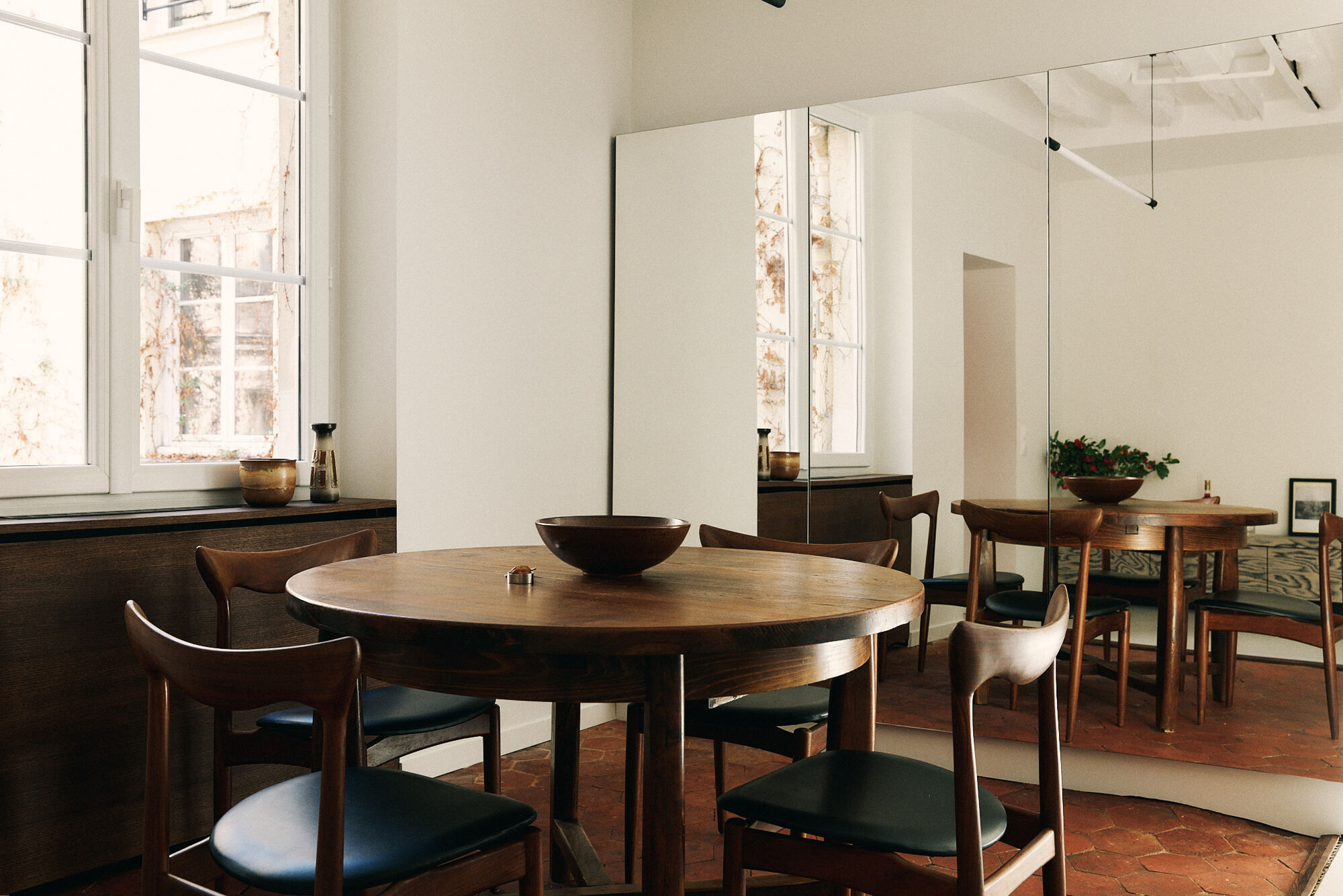 Round walnut dining table with leather-seated chairs on terracotta hex tiles, doubled in a full-height mirror beside a bright window