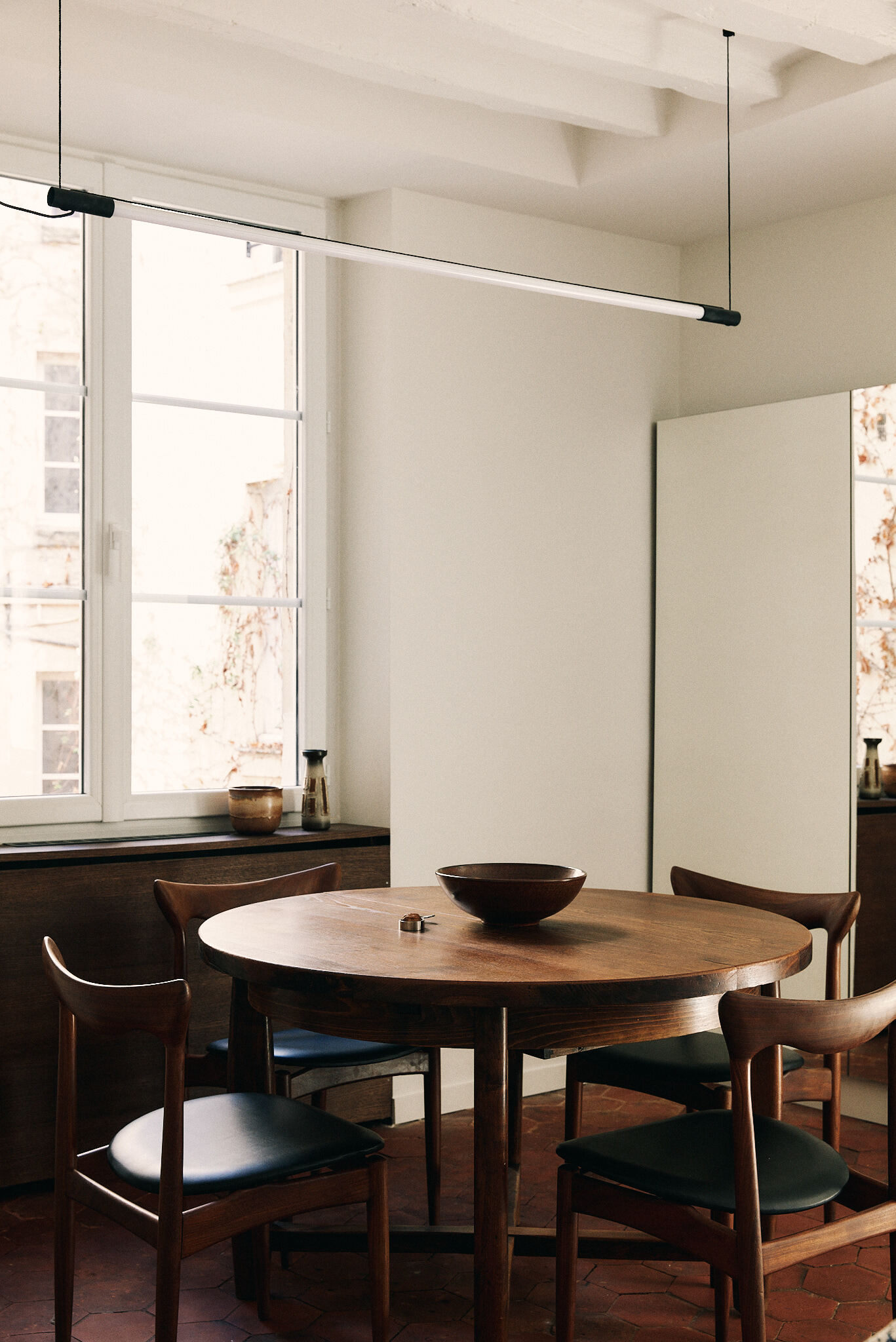 Round timber table and curved-back chairs beneath a slim linear pendant, with soft daylight through a tall white-framed window