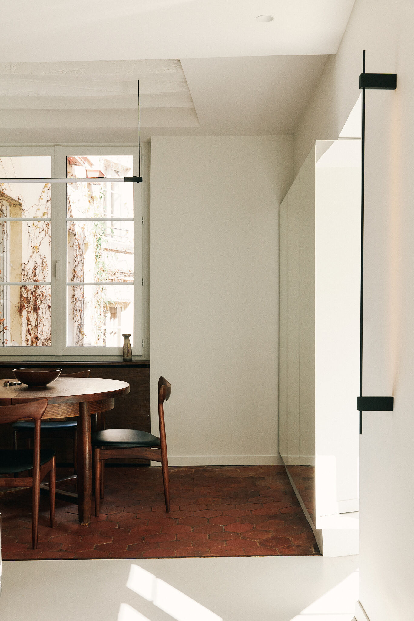 Terracotta tile floor meeting a poured pale floor, with a dining chair and a mirrored panel in low-angle sunlight