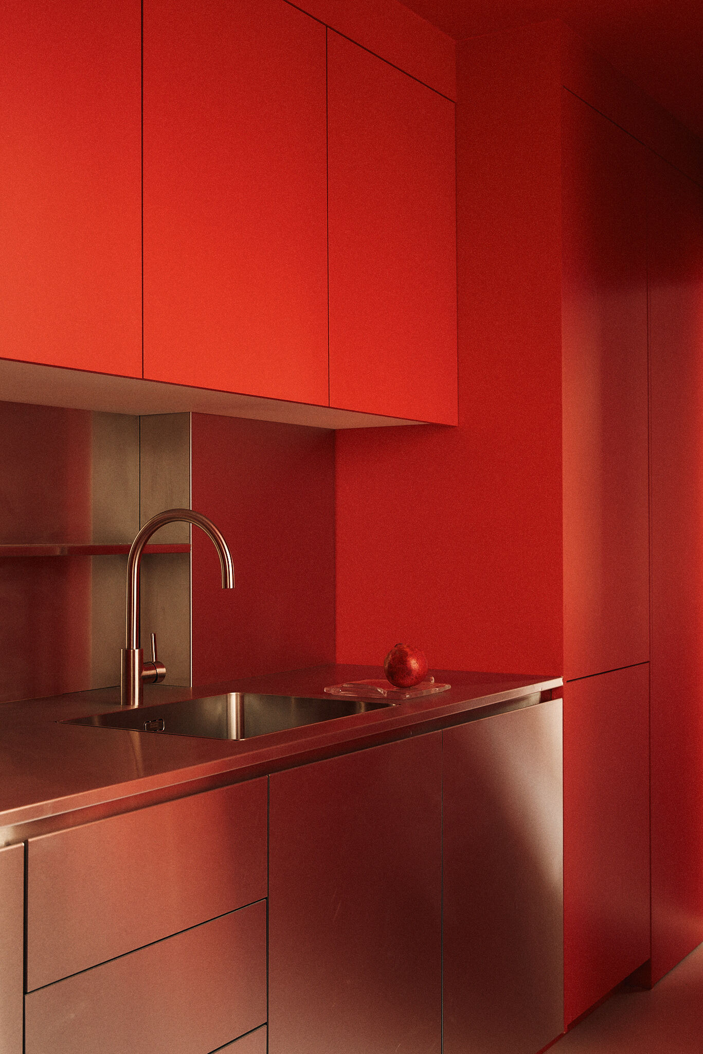 Red kitchen with flat cabinet fronts above a stainless steel counter, sink and curved tap, a pomegranate resting on the worktop