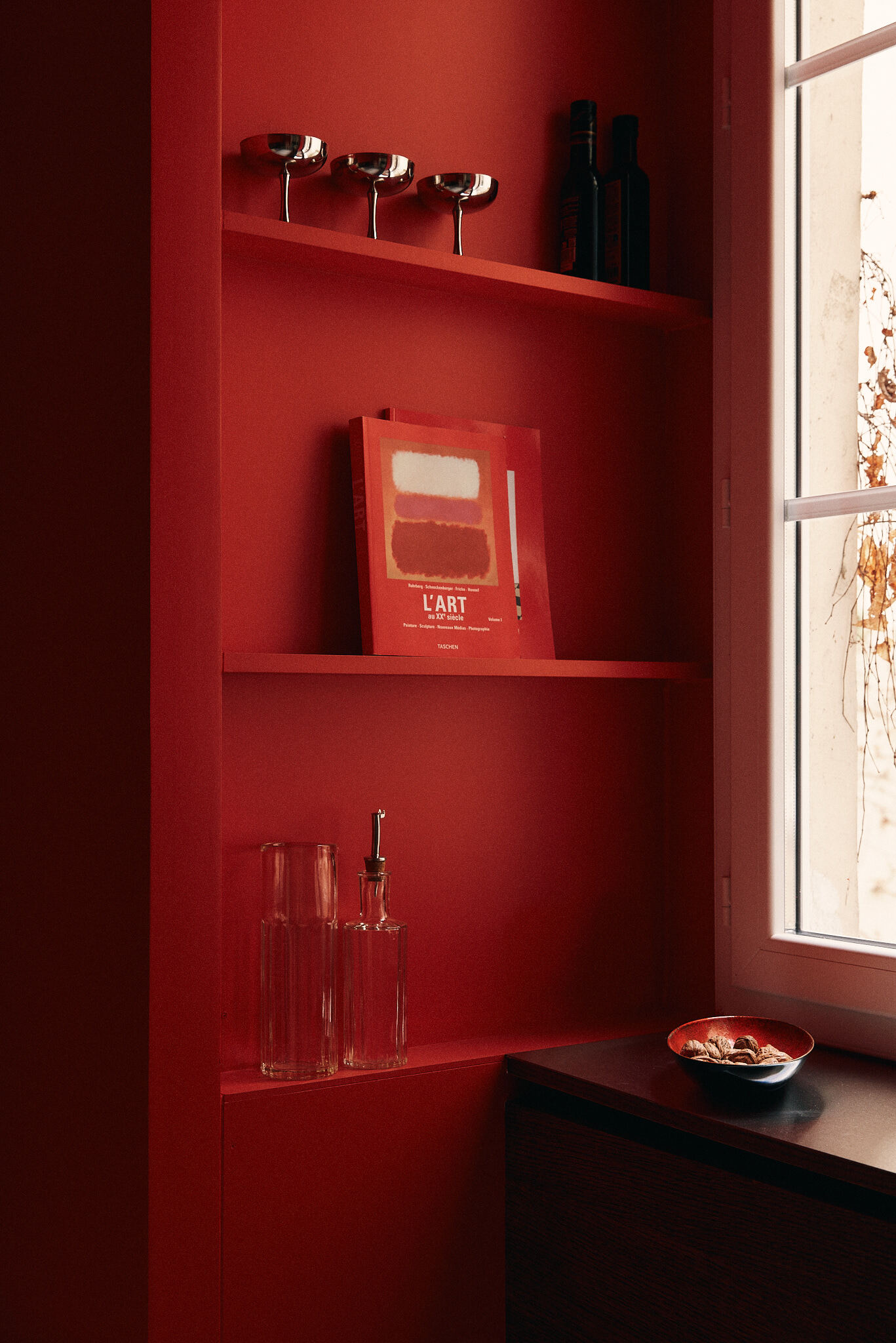 Red built-in shelving holding coupe glasses, an art book, carafes and bottles, beside a sunlit window with a bowl of nuts