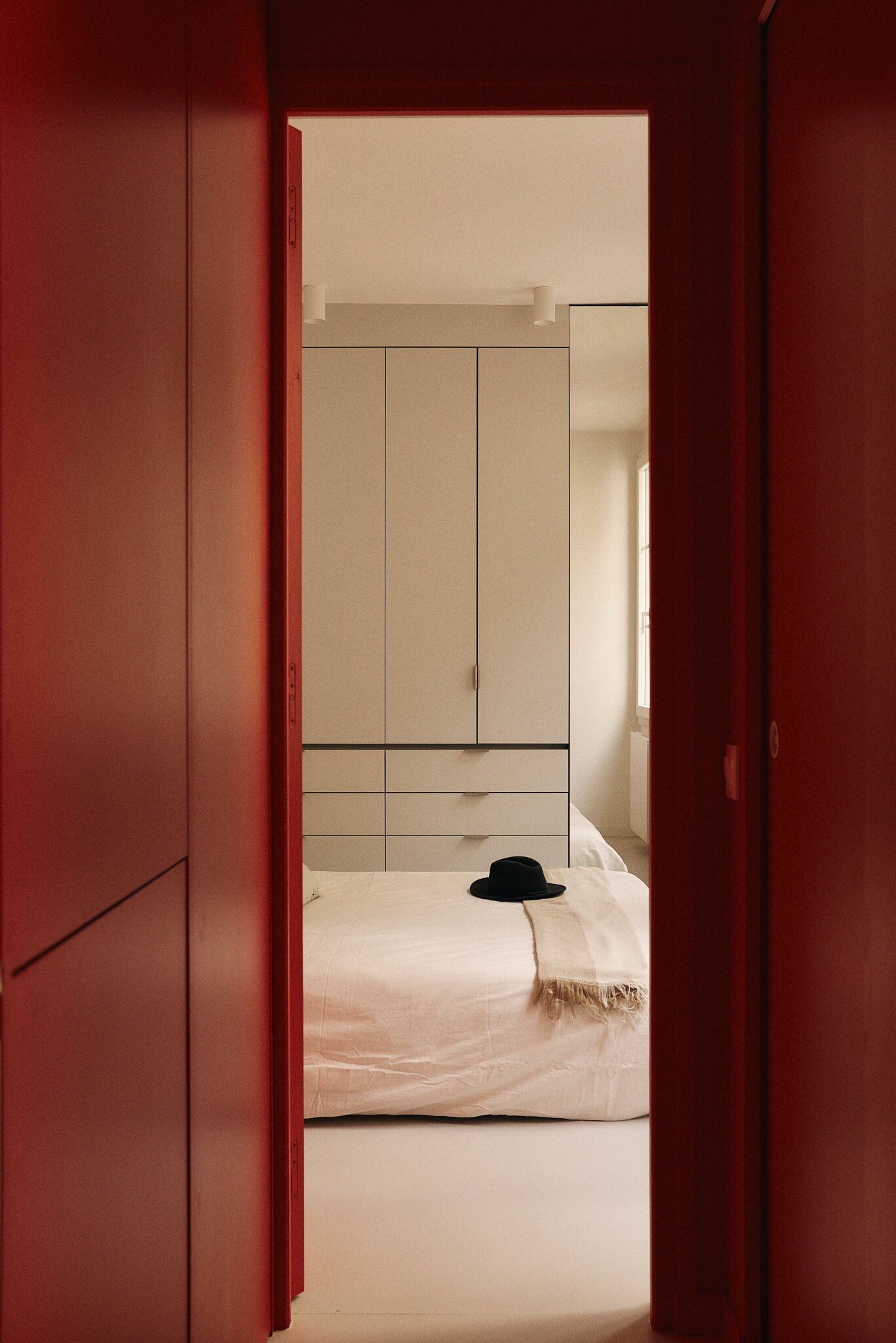 View through a red-painted doorway and paneled corridor to a bedroom with a white wardrobe and a black hat on the bed
