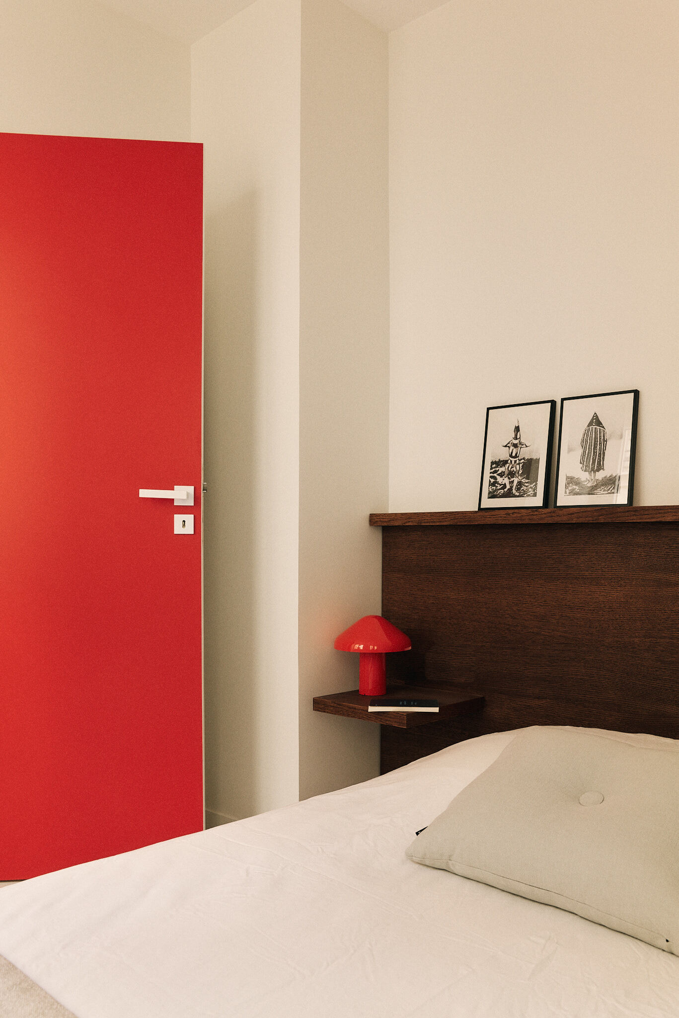 Red door beside a dark oak headboard with a shelf, framed prints and a small red mushroom lamp in soft daylight