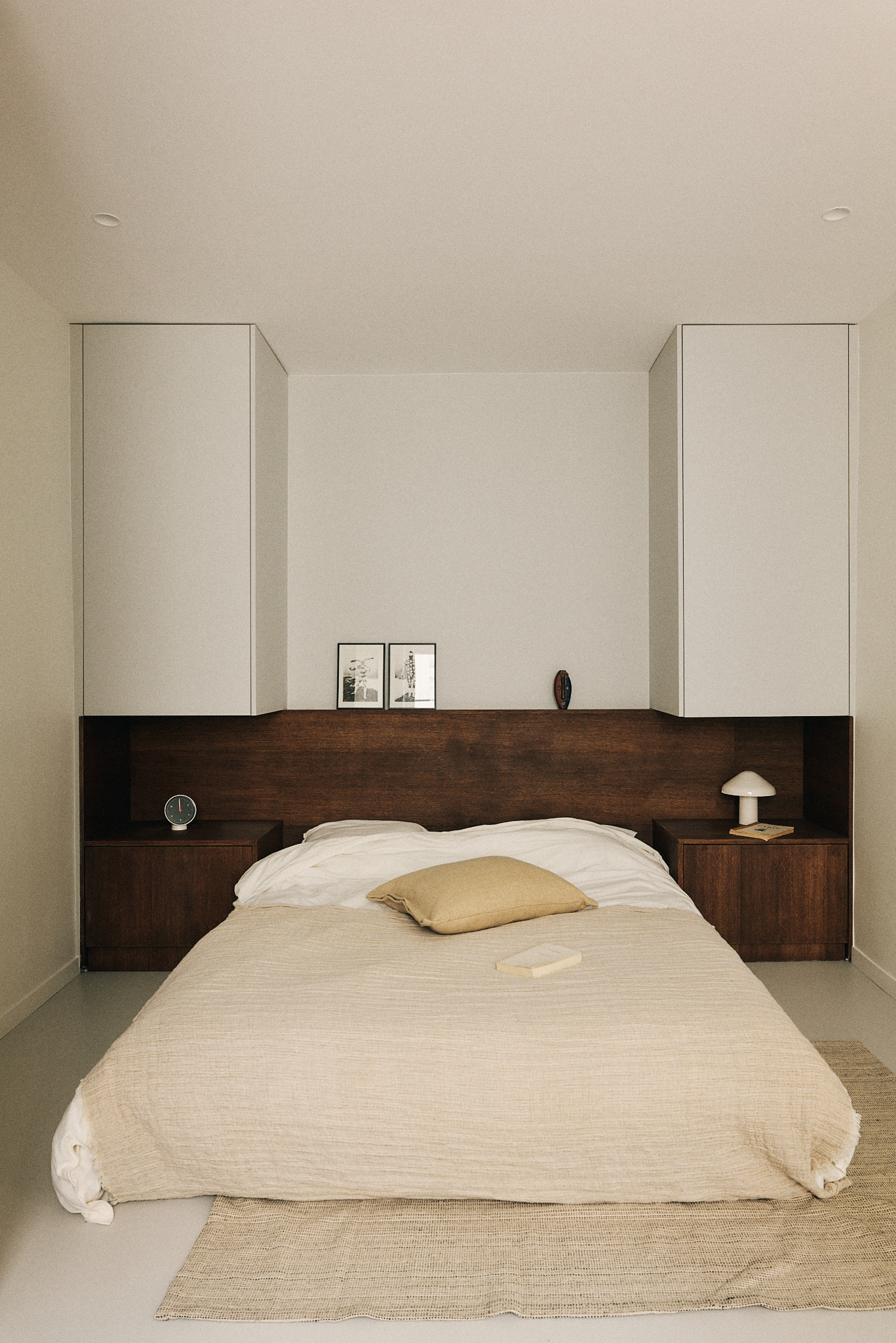 Symmetrical bed alcove with dark oak headboard, built-in nightstands and white overhead cabinets, a jute rug on the pale floor