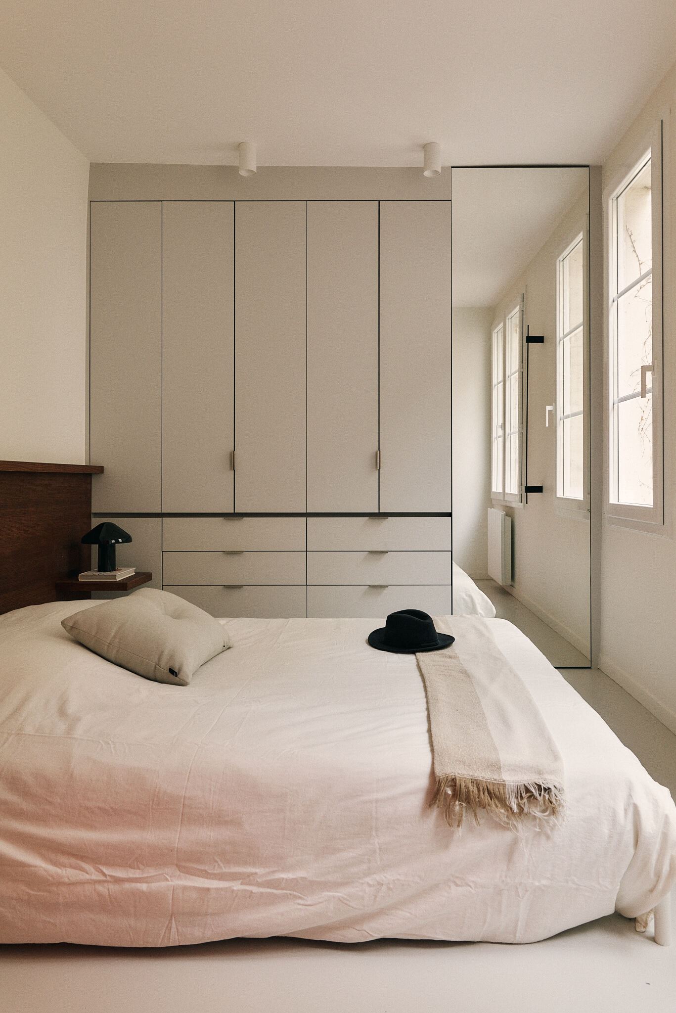 Bedroom with a full-height wardrobe, drawers and a tall mirror beside windows, a black hat resting on the linen bedding