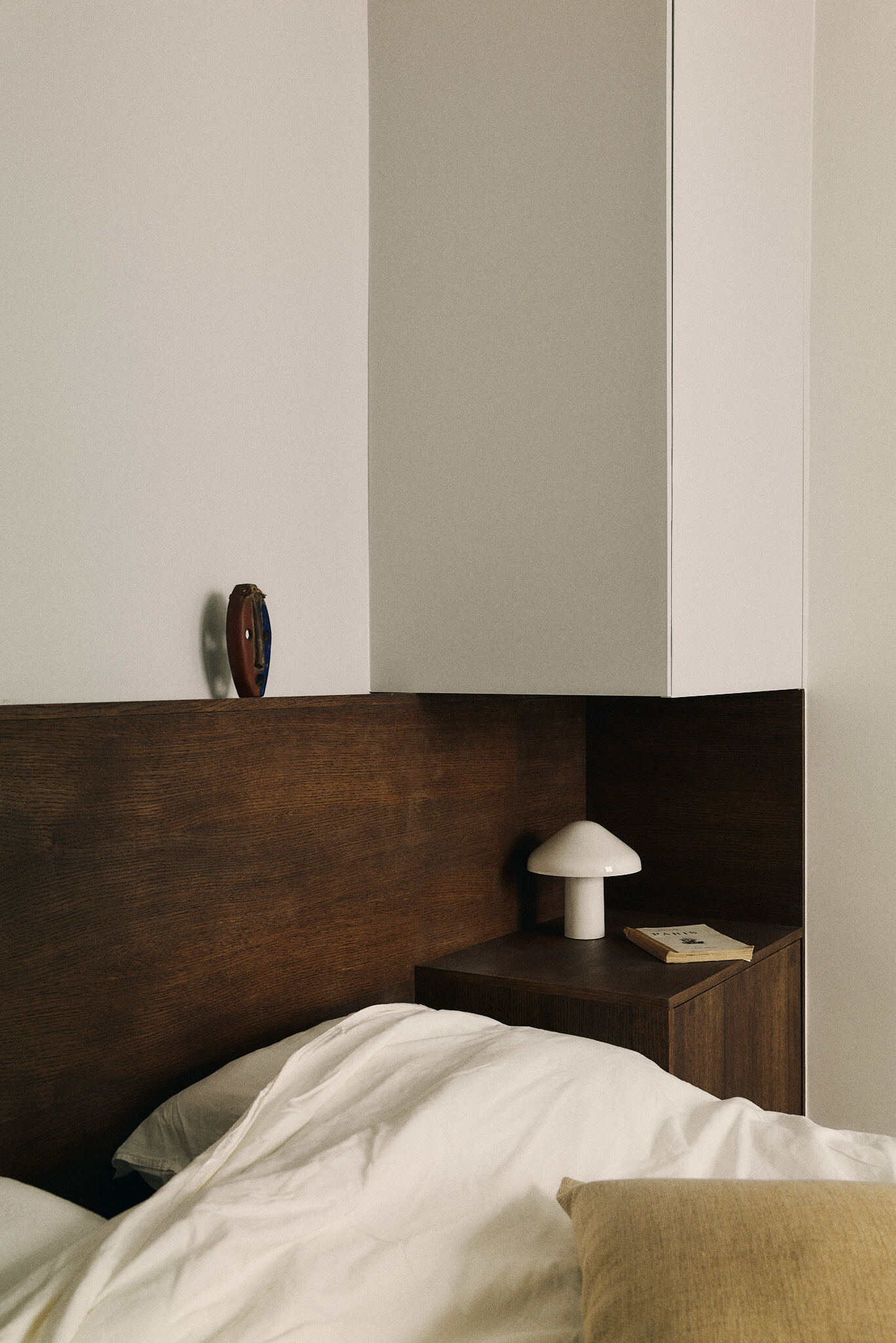 Close-up of the oak headboard, white overhead cabinet and a white mushroom lamp on the nightstand with a paperback