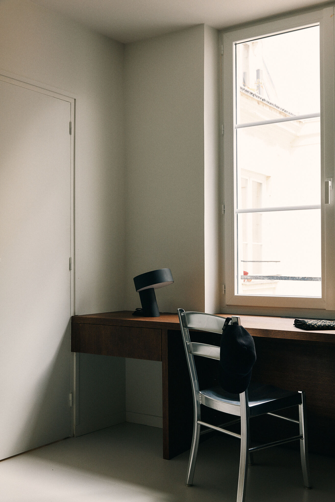 Timber desk beneath a tall window with a metal chair, a black lamp and a cap hanging on the backrest in diffuse daylight