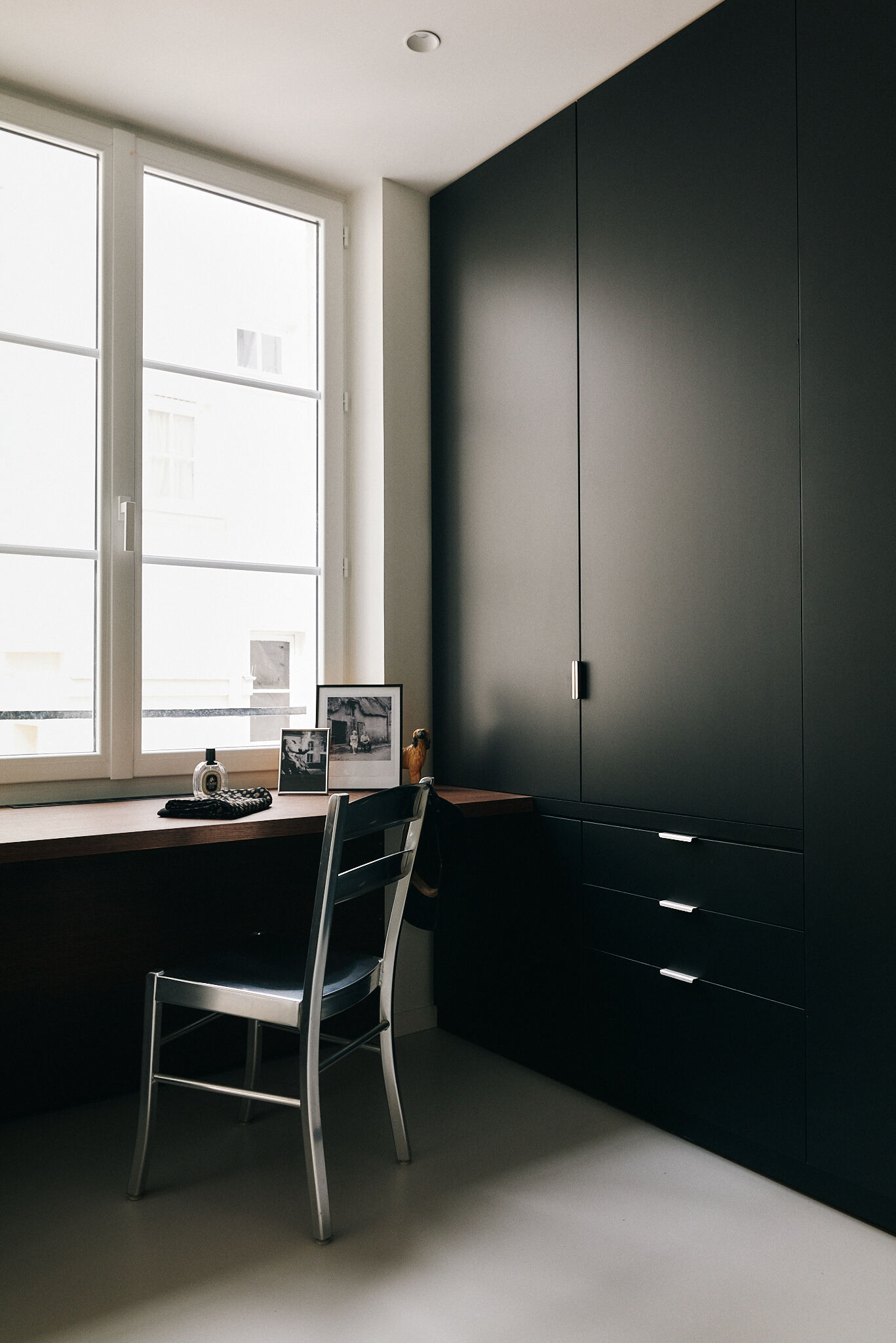 Built-in desk beneath a tall casement window, with a metal chair and floor-to-ceiling black cabinets in soft daylight