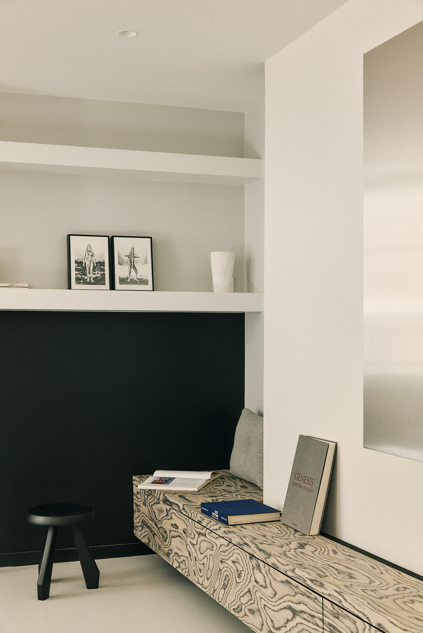 Window-side nook with a swirled-grain wood bench, stacked books and a black stool beneath a white shelf of framed prints