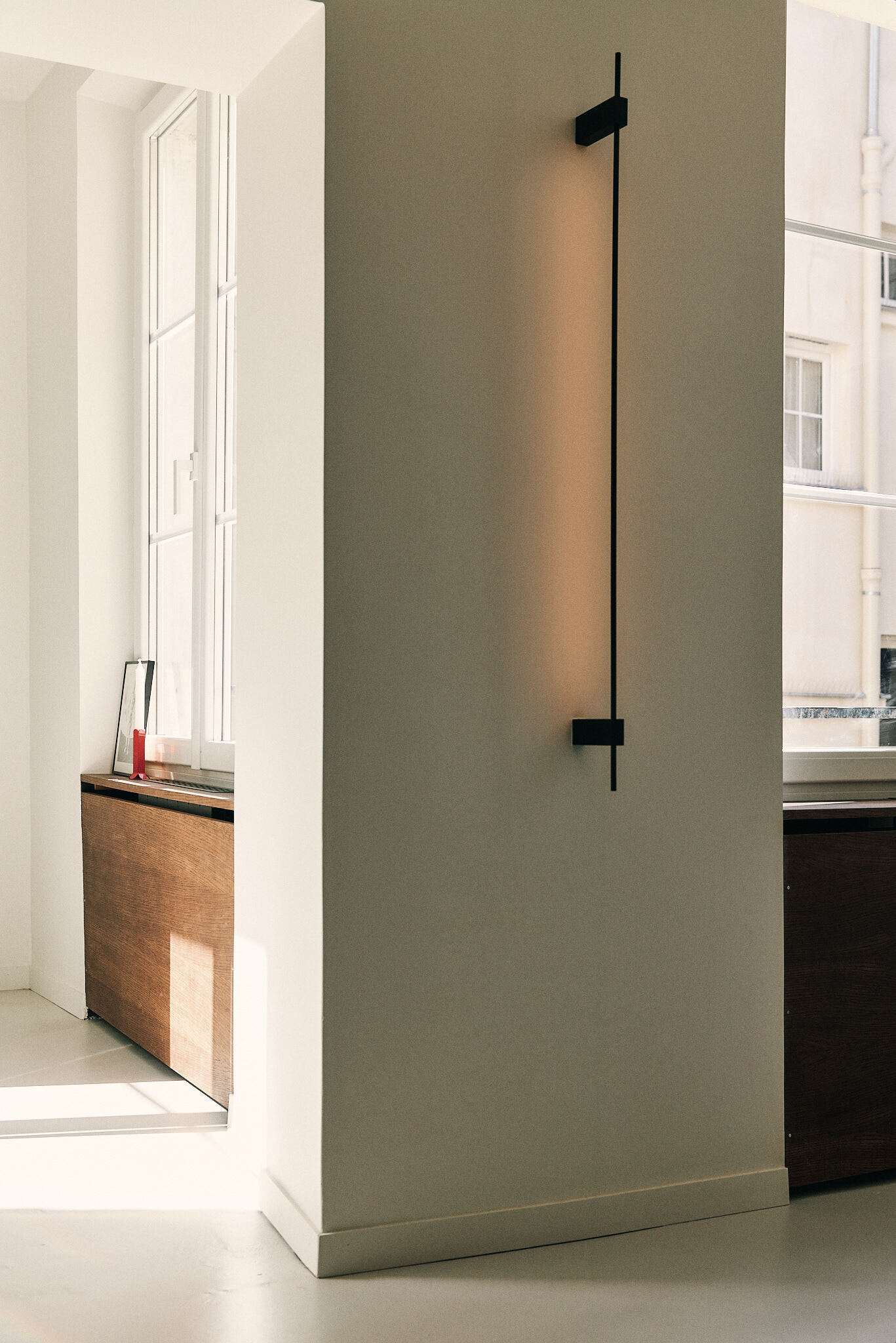 Slim vertical black wall light glowing on a white corner column, with a sunlit window and oak radiator cover beyond