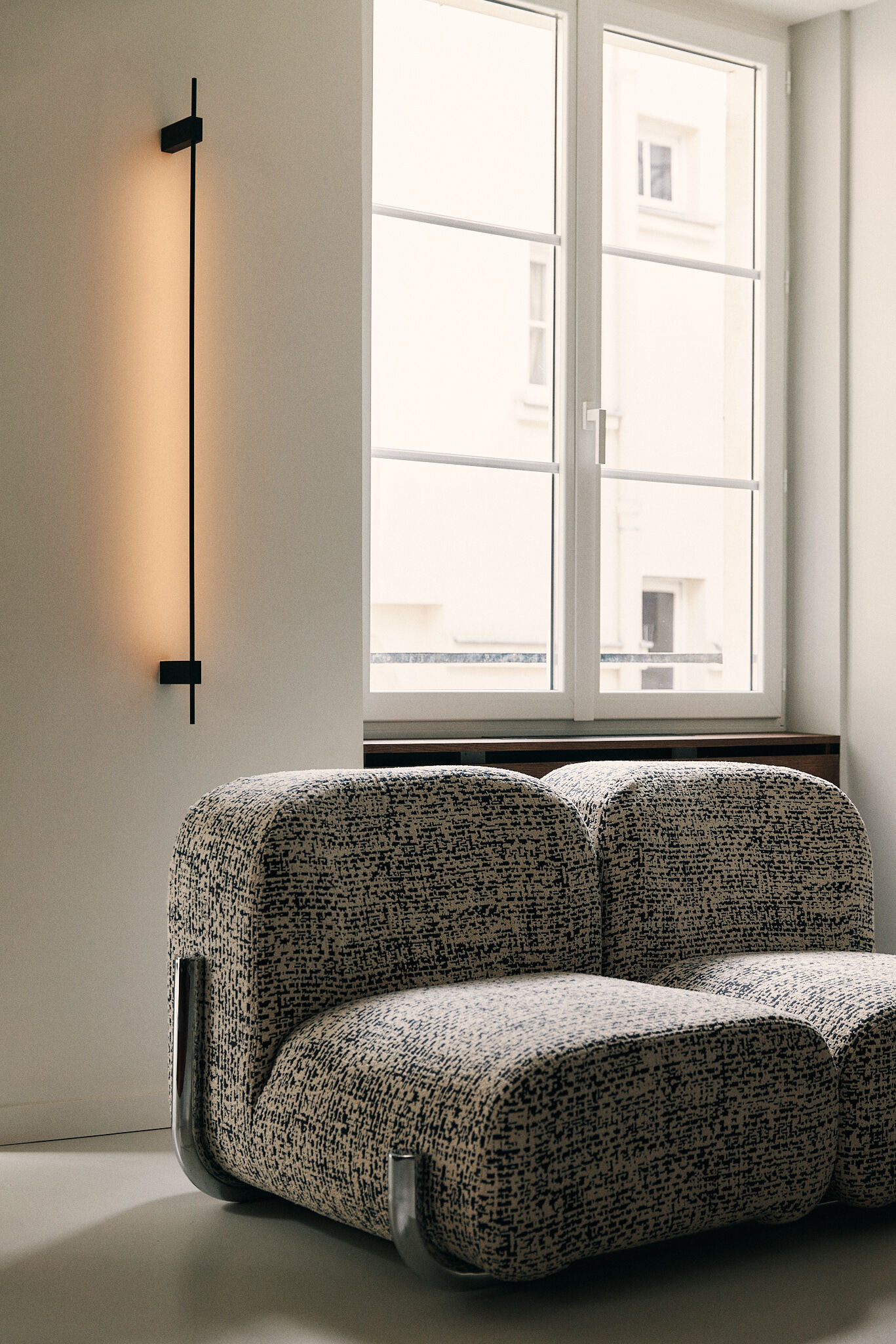 Bouclé modular armchair with chrome feet beneath a tall casement window and a slim vertical wall light glowing in soft daylight