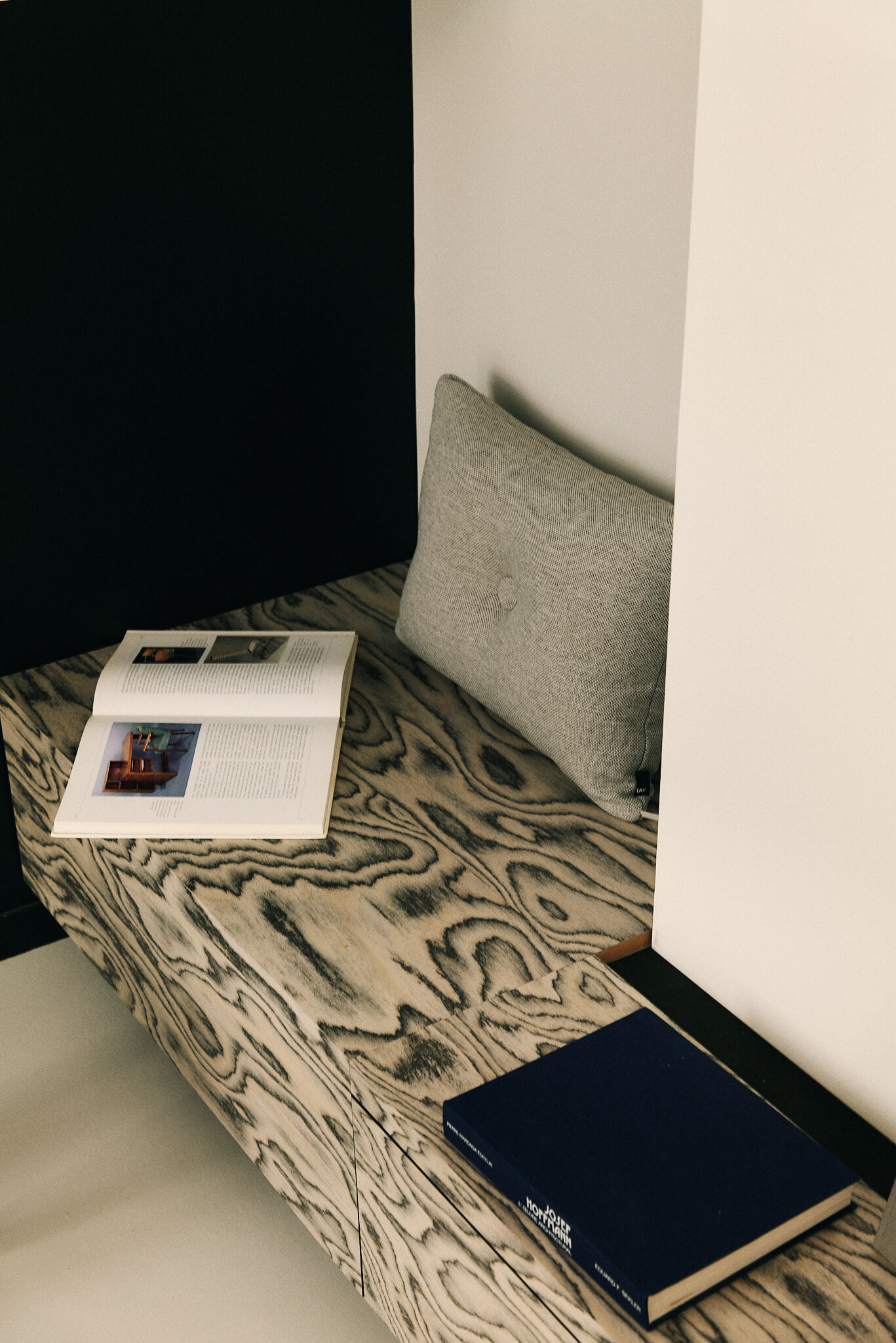 Built-in bench of grain-patterned timber with a grey cushion, an open book and a navy hardback beside a dark wall