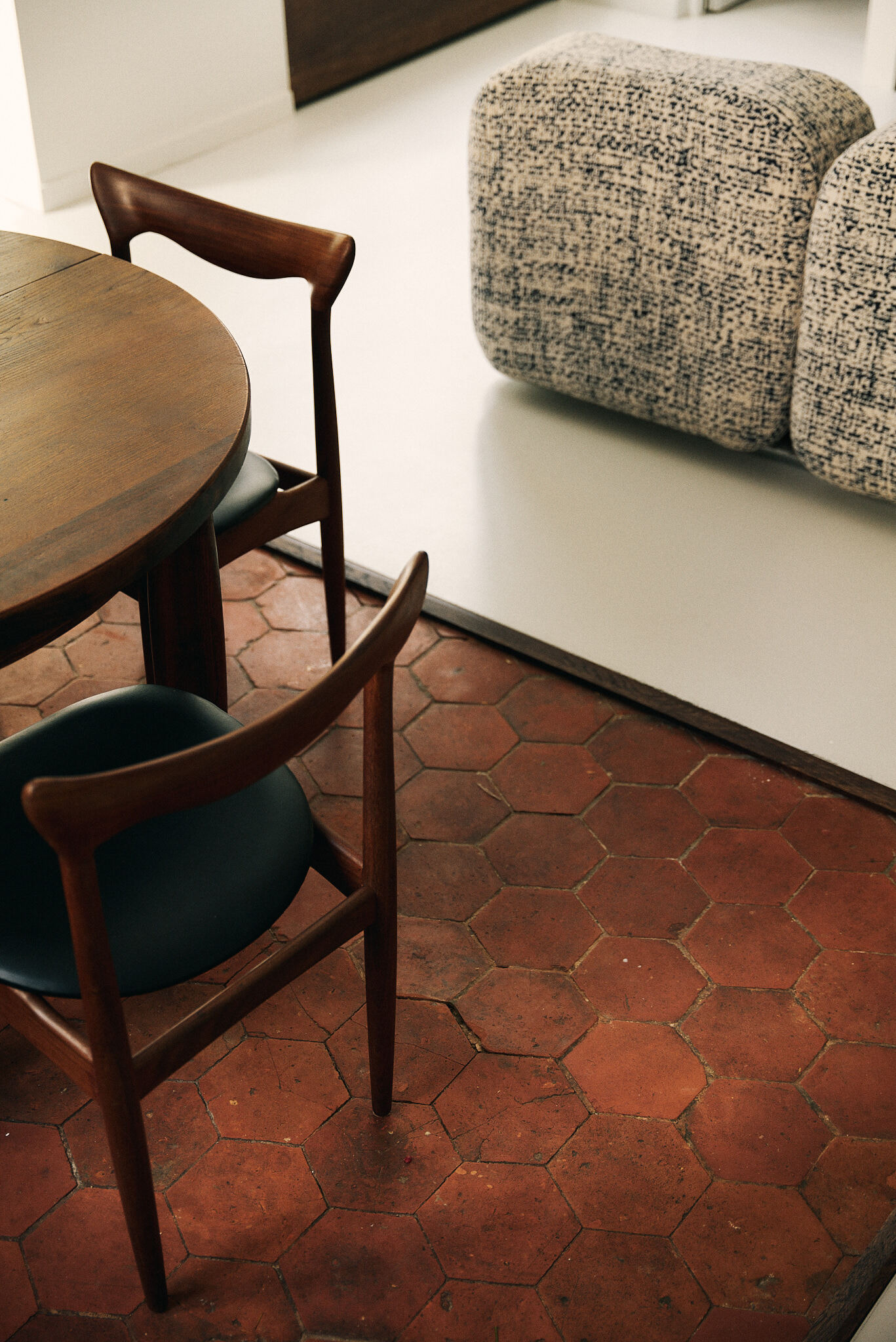 Round timber dining table and teak chairs on hexagonal terracotta tiles, with a raised pale floor and bouclé seating beyond