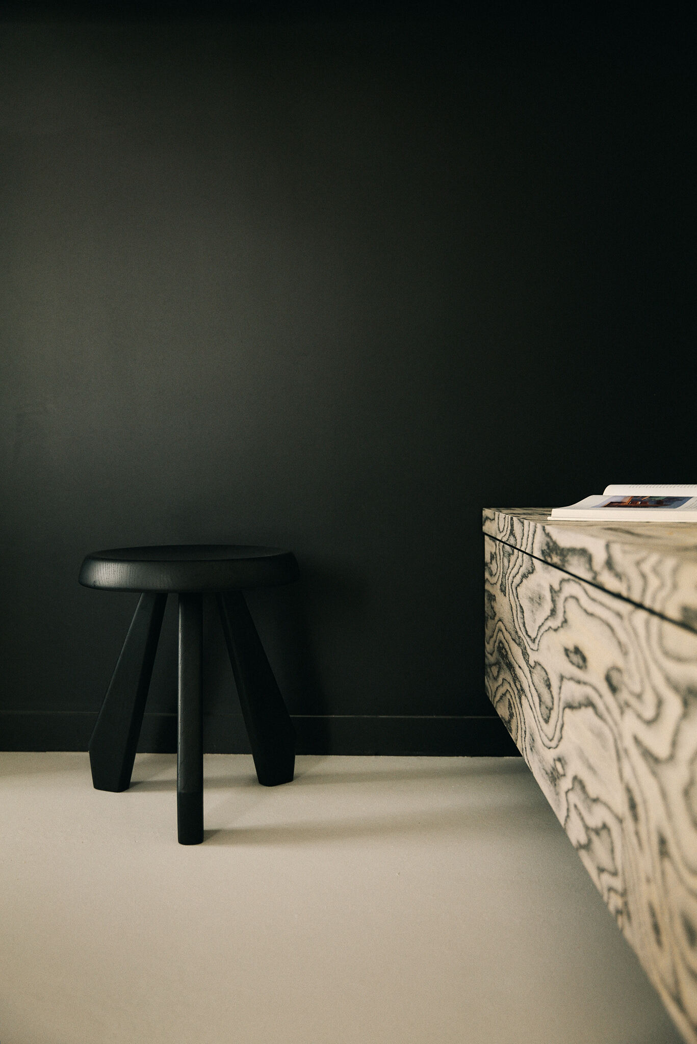 Black three-legged wooden stool against a dark painted wall beside the edge of a floating bench with bold timber grain