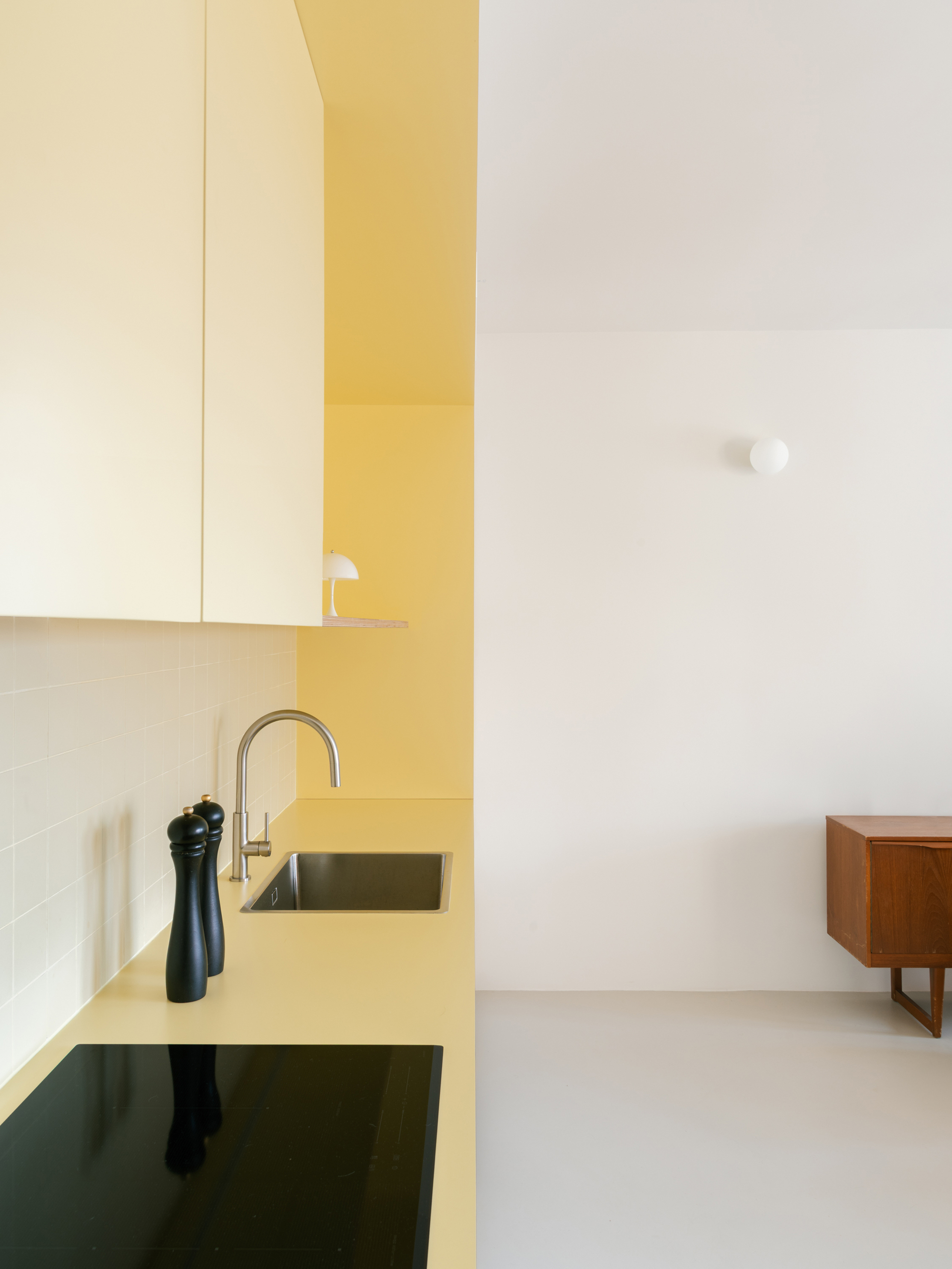 Side view of the yellow countertop with sink, induction hob and two black pepper mills, a teak sideboard beyond