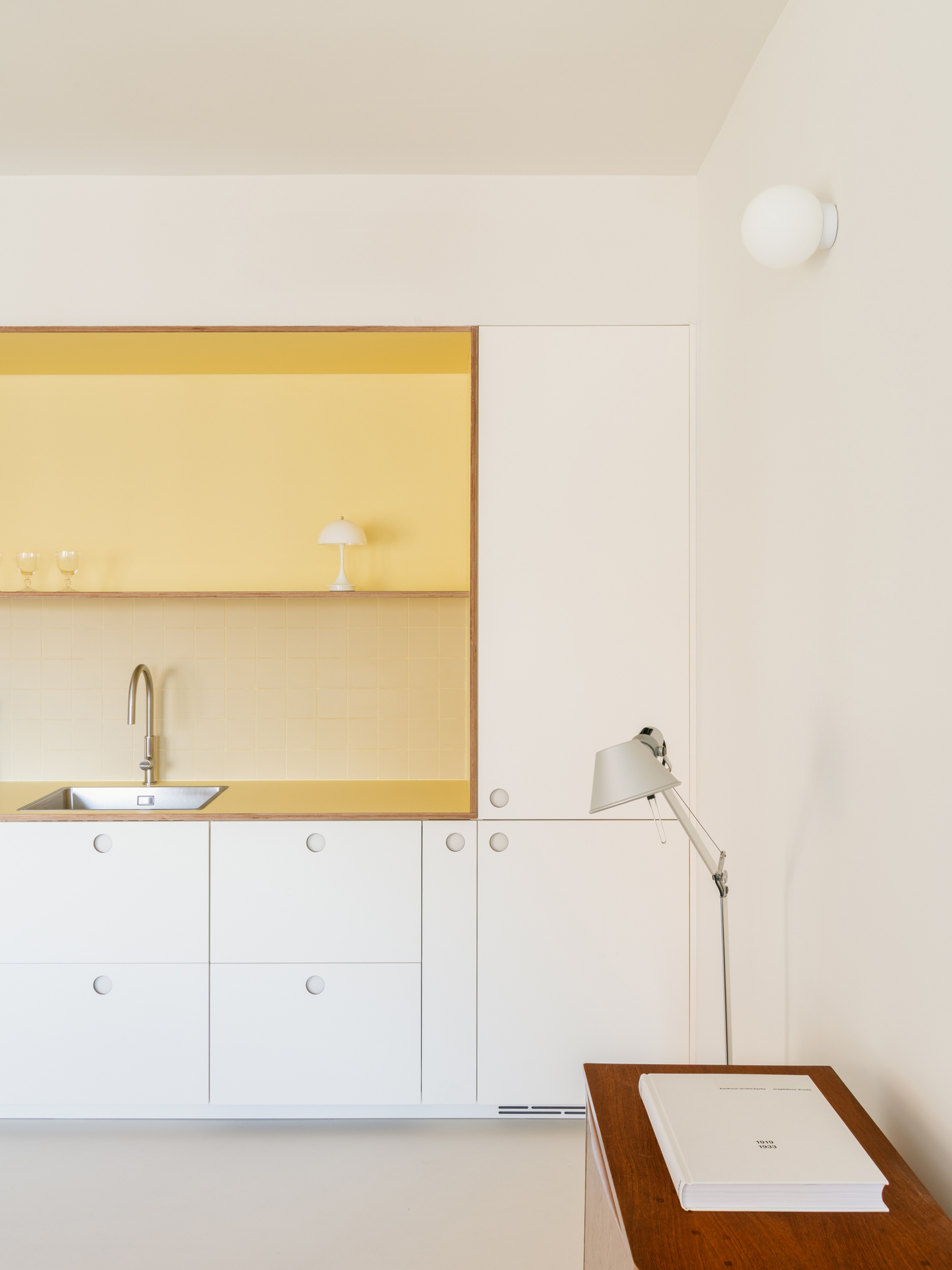 Yellow kitchen niche framed in plywood above white drawers, with an articulated desk lamp and a book on a teak table