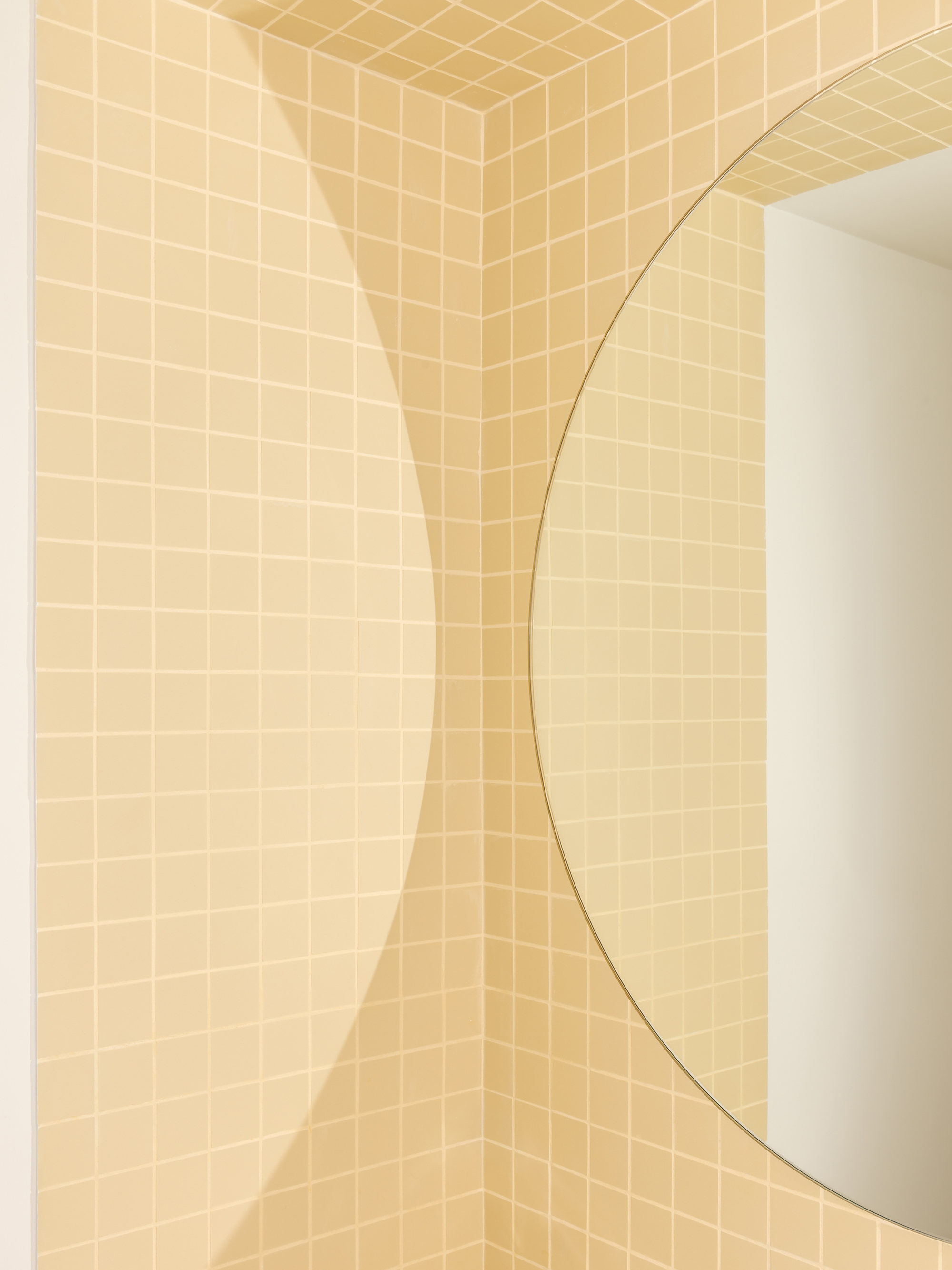 Corner of square cream tiles meeting a round mirror, with a soft curved shadow falling across the wall