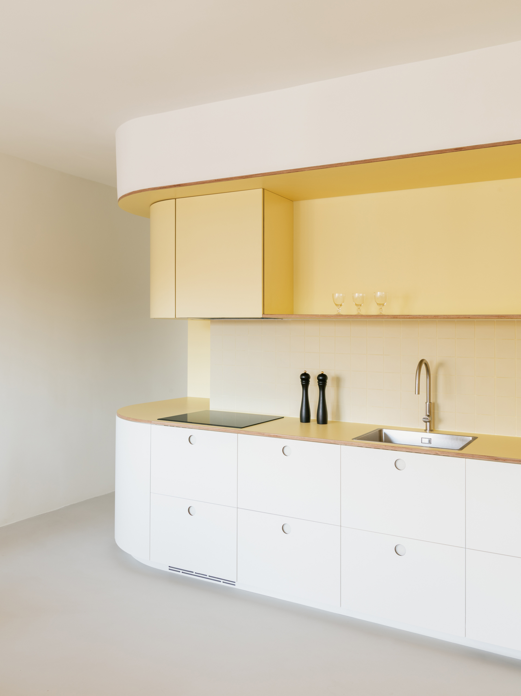 Angled view of the curved white kitchen run with a yellow overhead cabinet, wine glasses on the shelf and daylight from the left