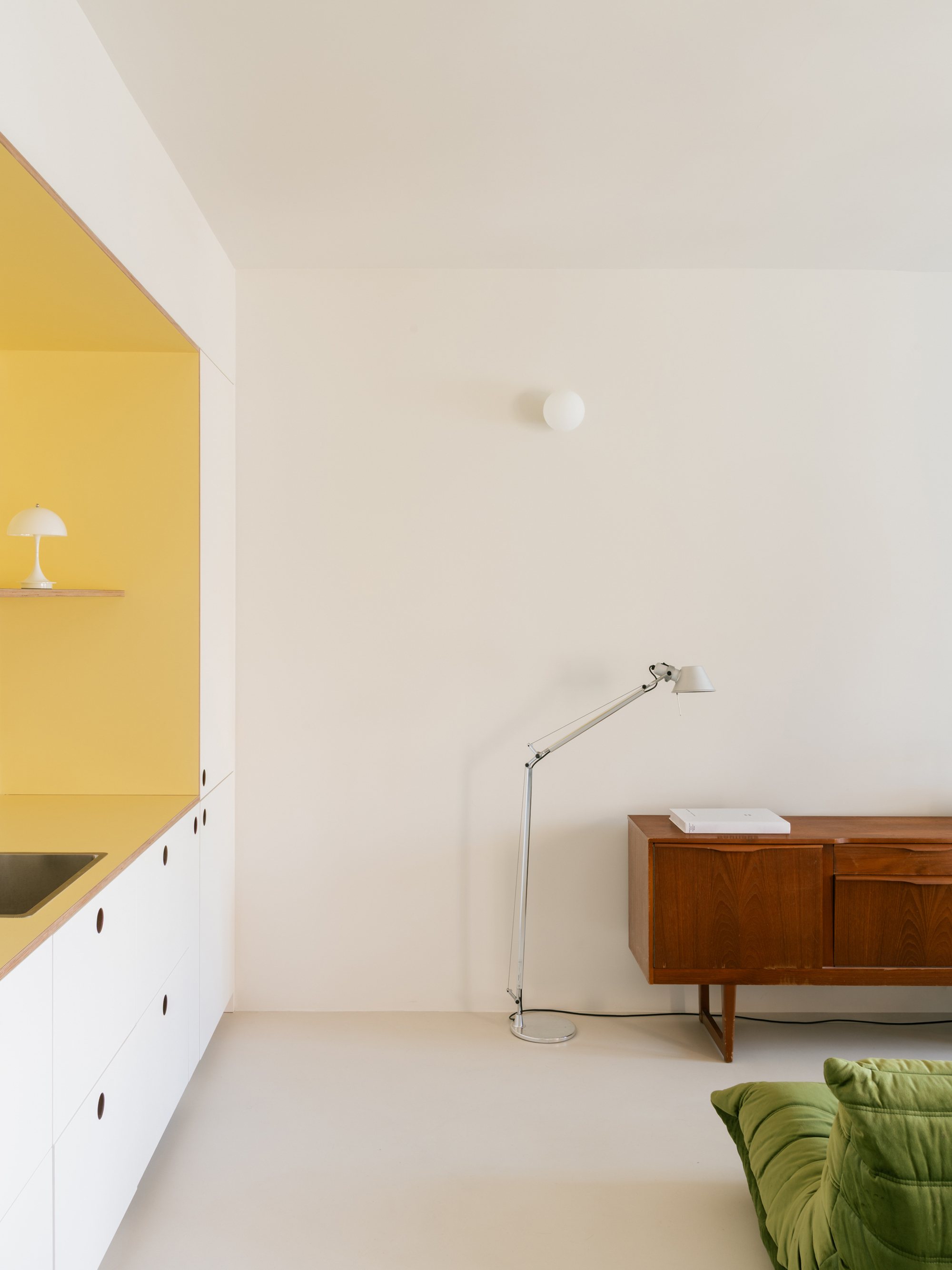 Yellow-lined kitchen niche beside a floor lamp, teak sideboard and green quilted sofa in soft daylight