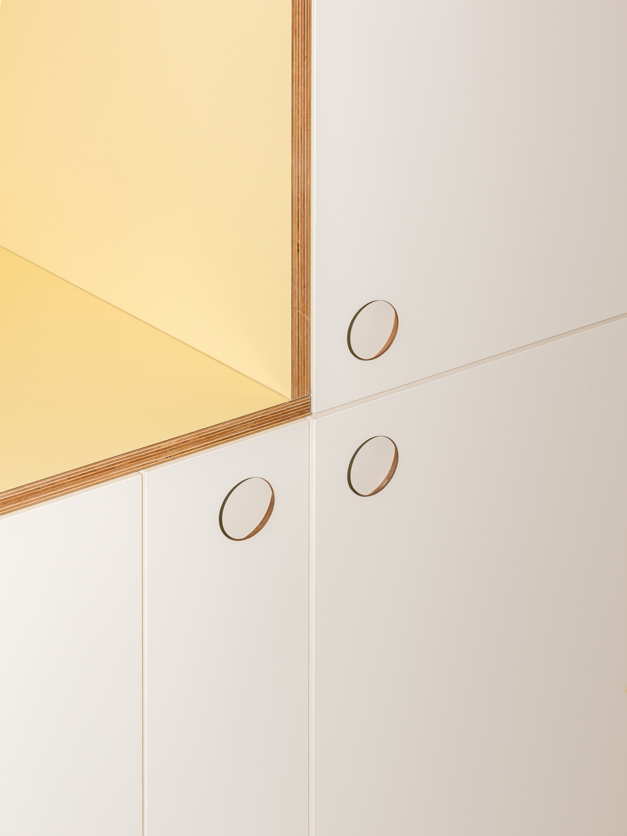 Corner detail of white cabinet doors with circular finger pulls beside a yellow niche edged in plywood