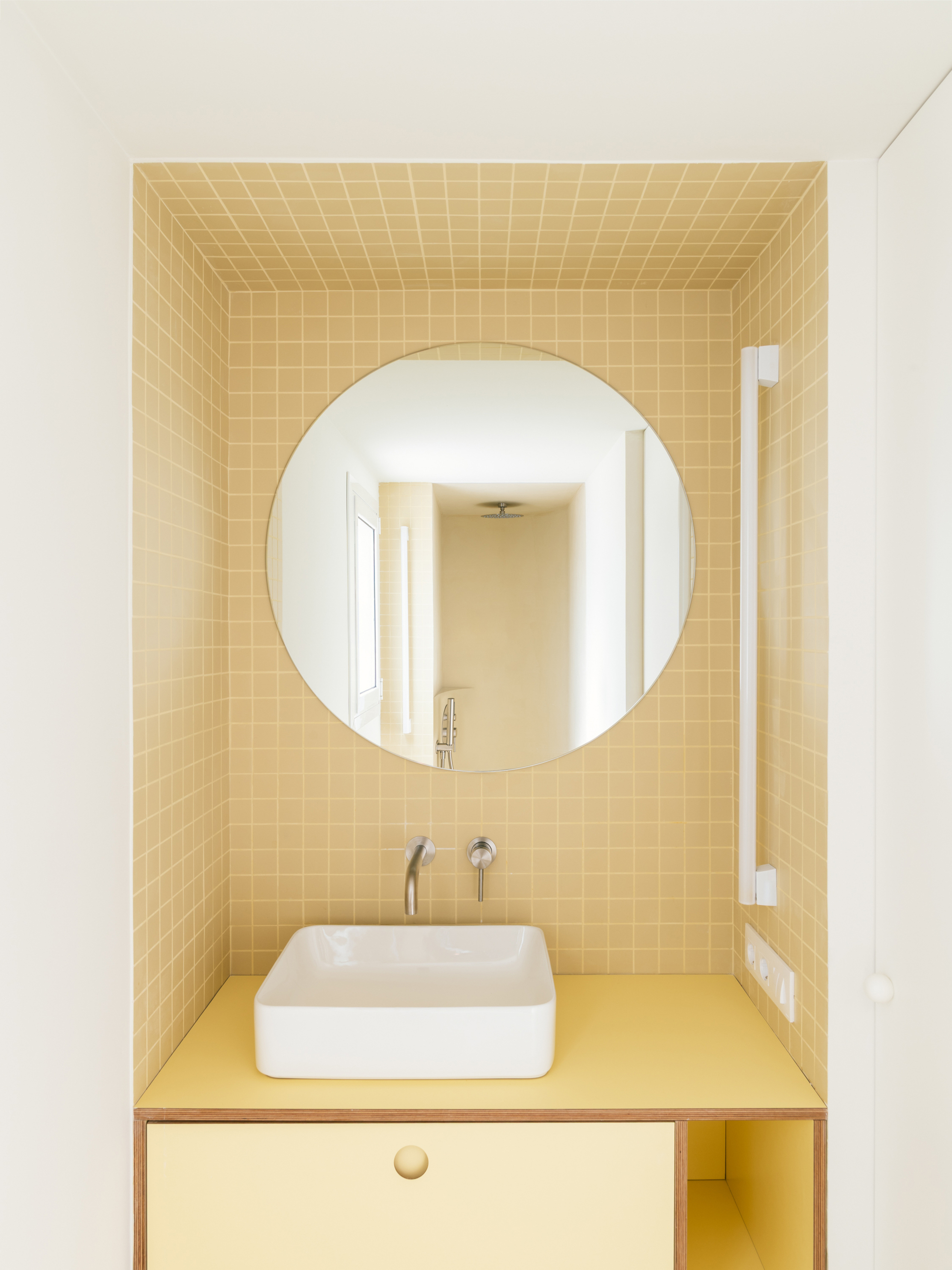 Tiled yellow bathroom alcove with round mirror, wall-mounted tap, white basin and yellow plywood vanity in even daylight