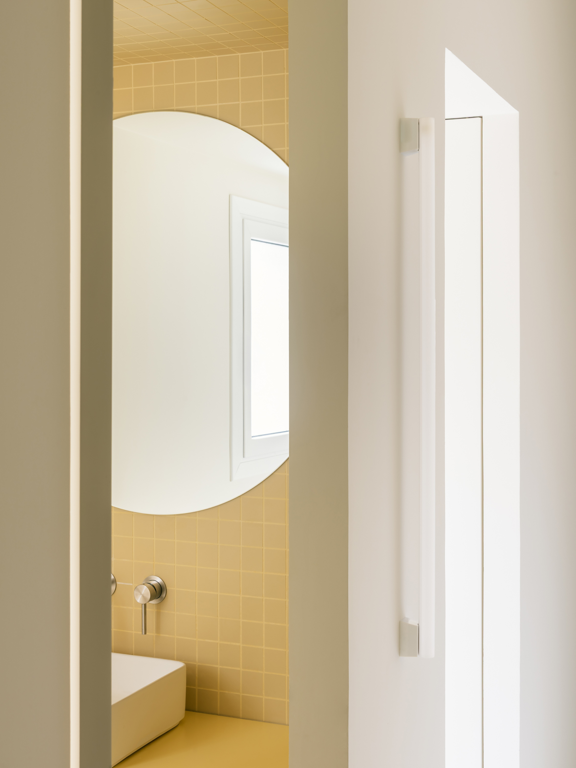 Half-open door revealing a round mirror, window reflection and square yellow tiles beside a long white handle