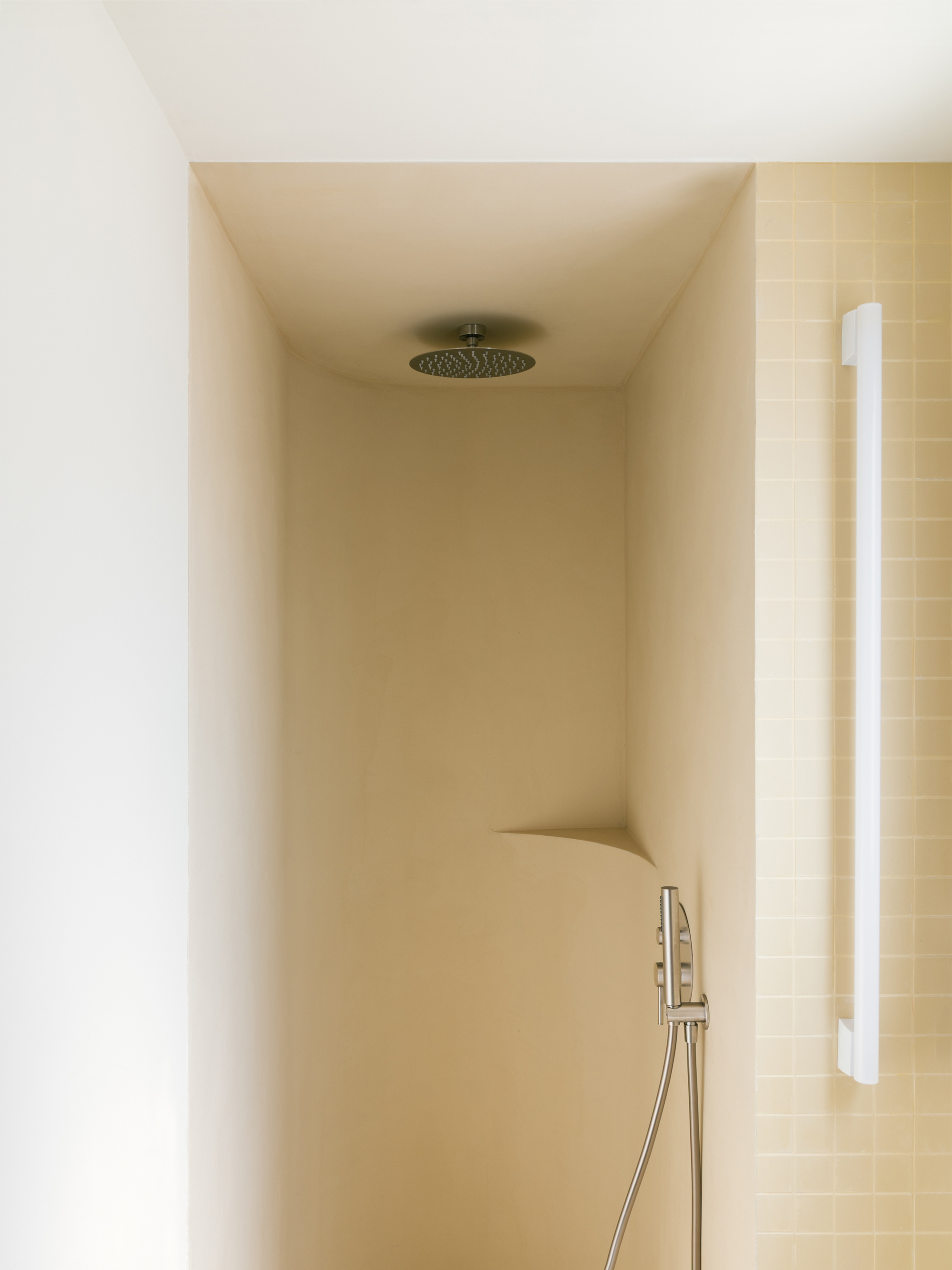 Shower alcove with curved ochre plaster walls, a round ceiling rain head and a white grab bar against small square tiles in soft daylight