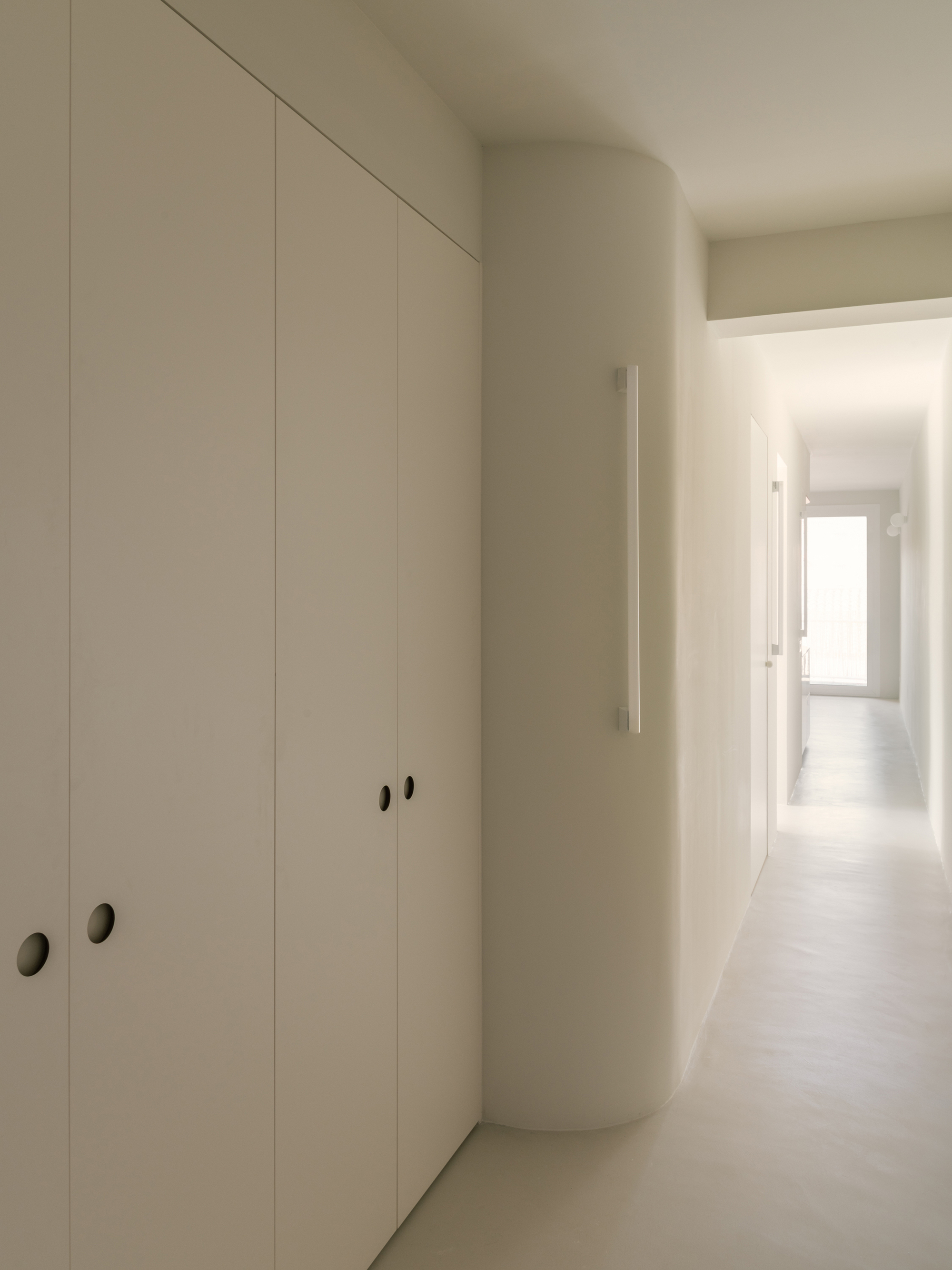 Hallway with flush white wardrobe doors and a rounded wall corner leading toward a bright window at the far end