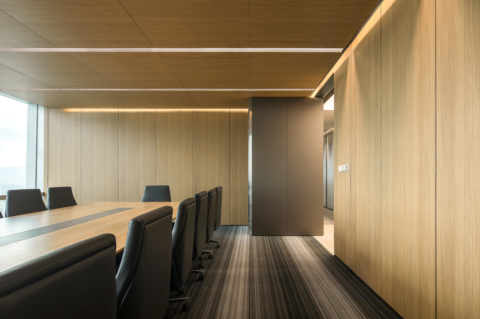 Conference room corridor edge with oak veneer wall panels, a bronze-toned freestanding core and striped carpet beside black leather chairs in daylight