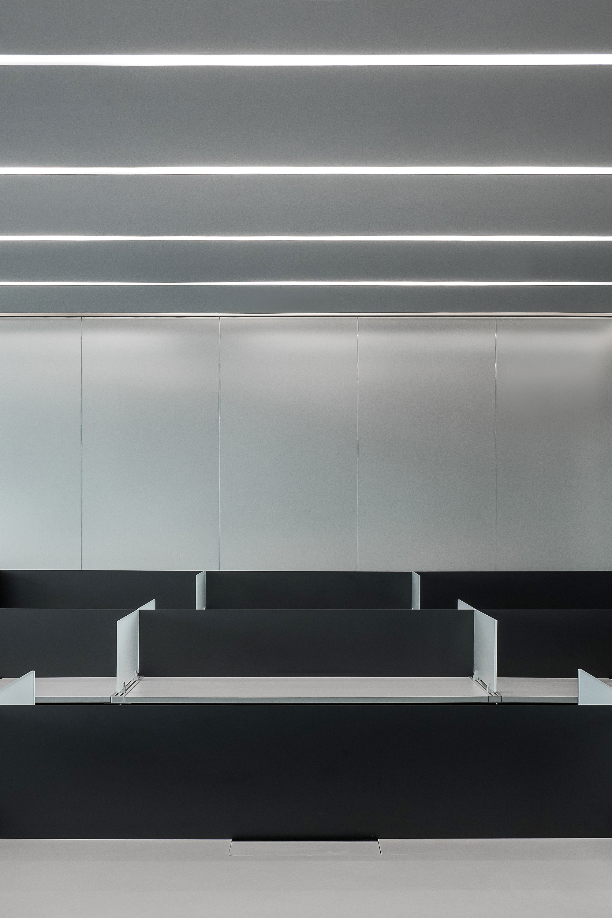 Black partitioned desk bays in front of a pale metal panel wall, under a ceiling of parallel linear light strips