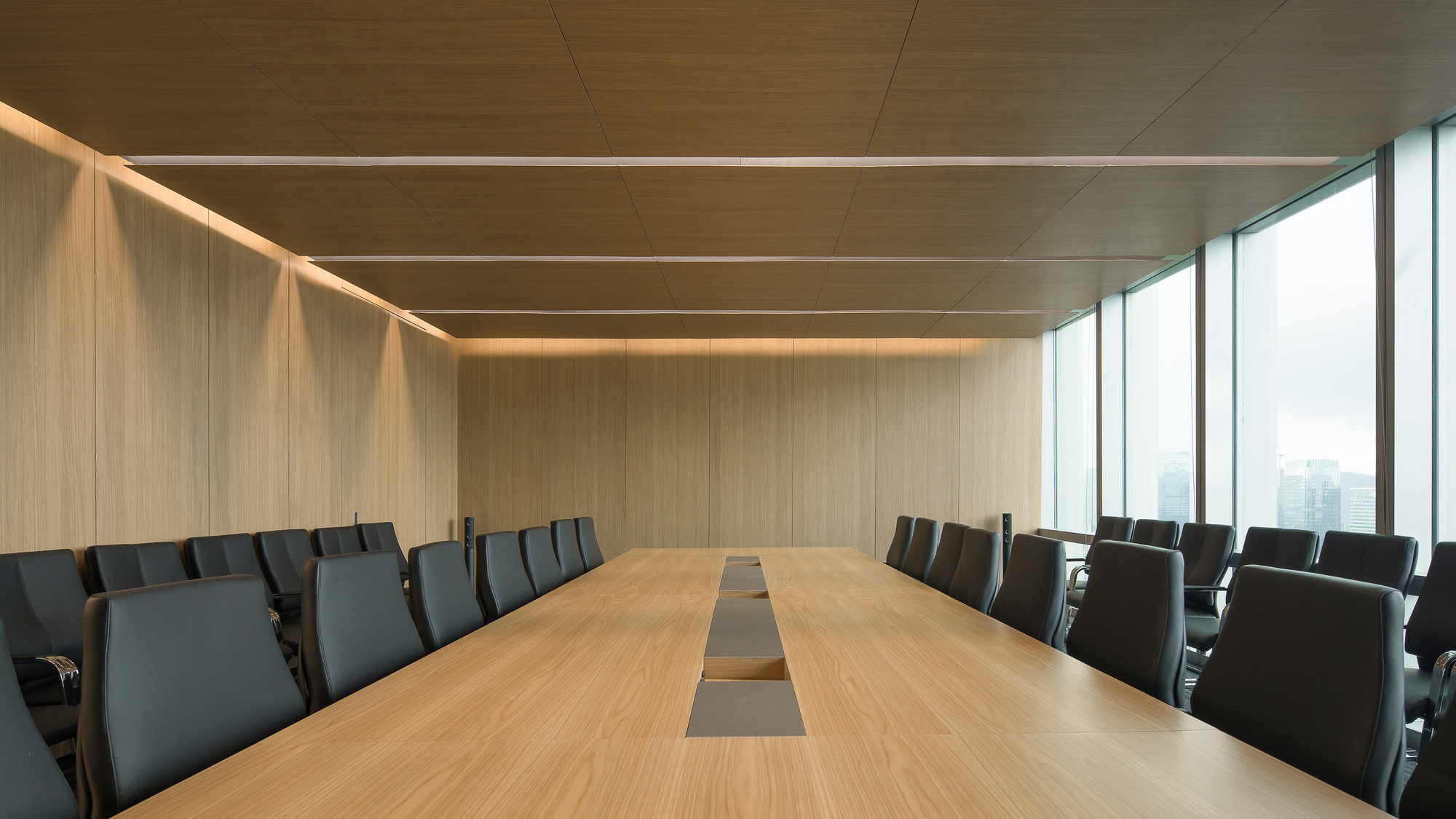 Long oak meeting table with recessed central trough, flanked by black chairs and a floor-to-ceiling window opening onto a hazy skyline