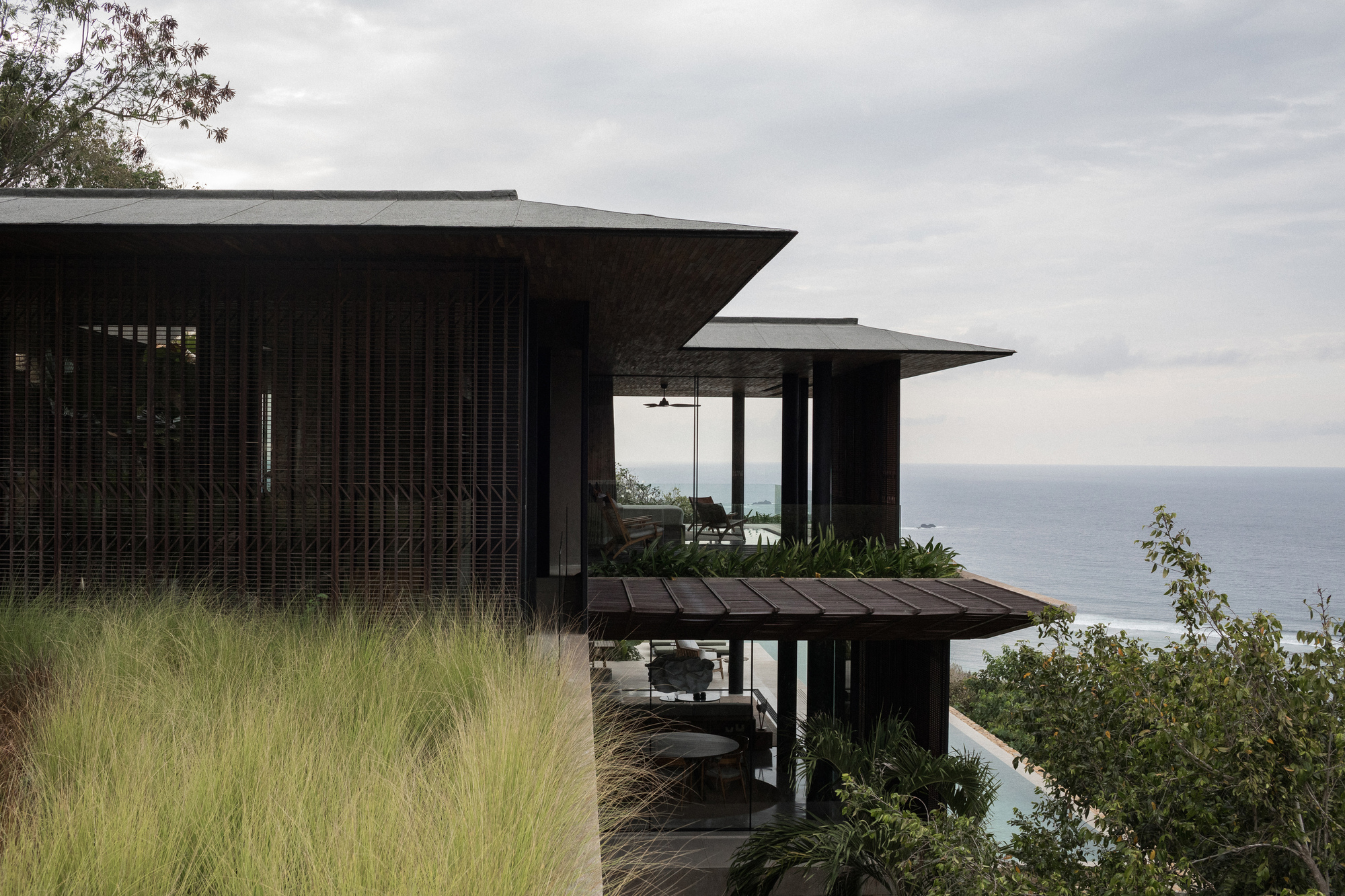 Slatted timber screen wall and overhanging dark roofs above a grass terrace, with the ocean beyond under an overcast sky