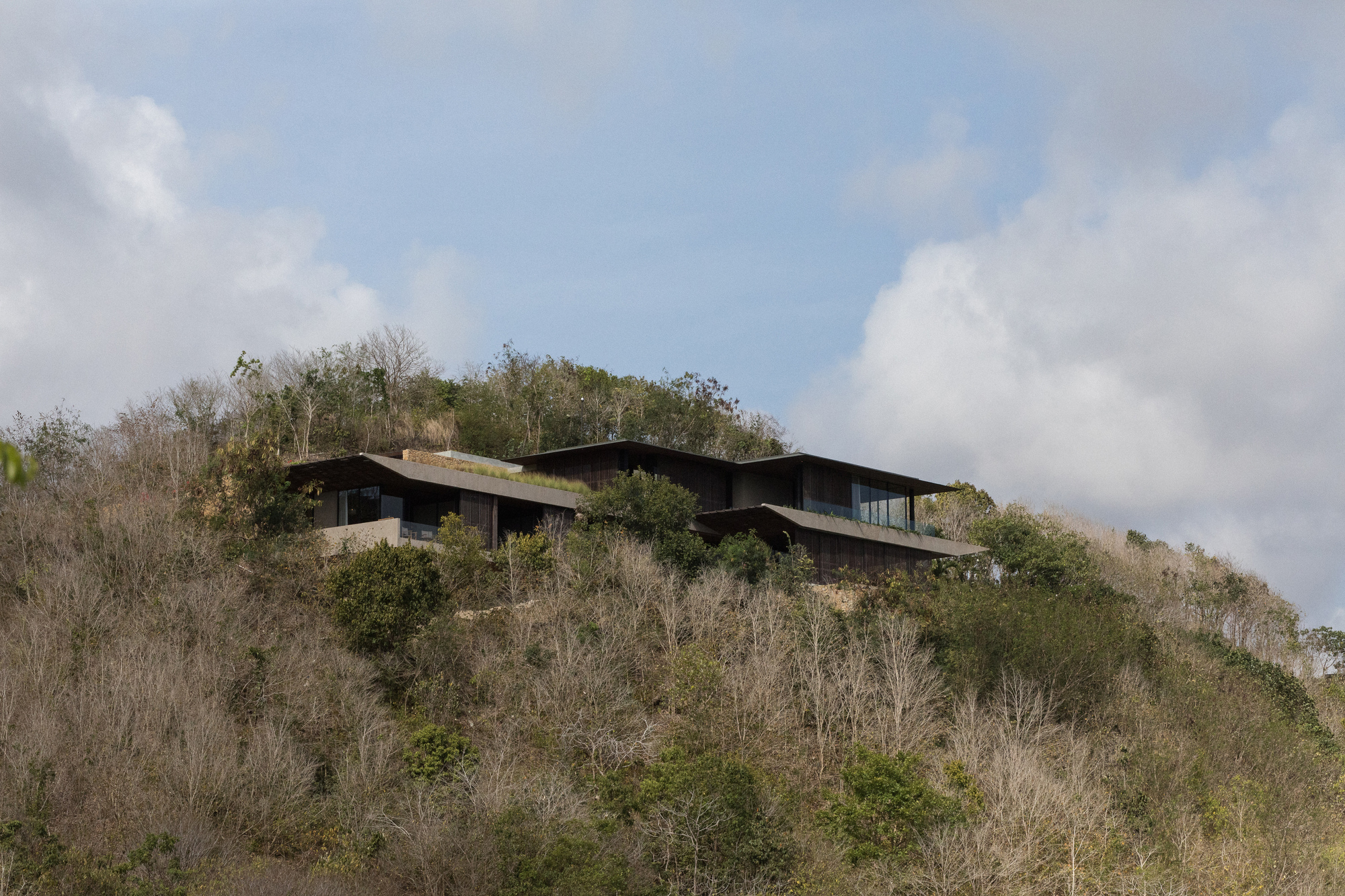 Distant view of the low house with broad eaves perched on a dry scrubby hillside under towering clouds