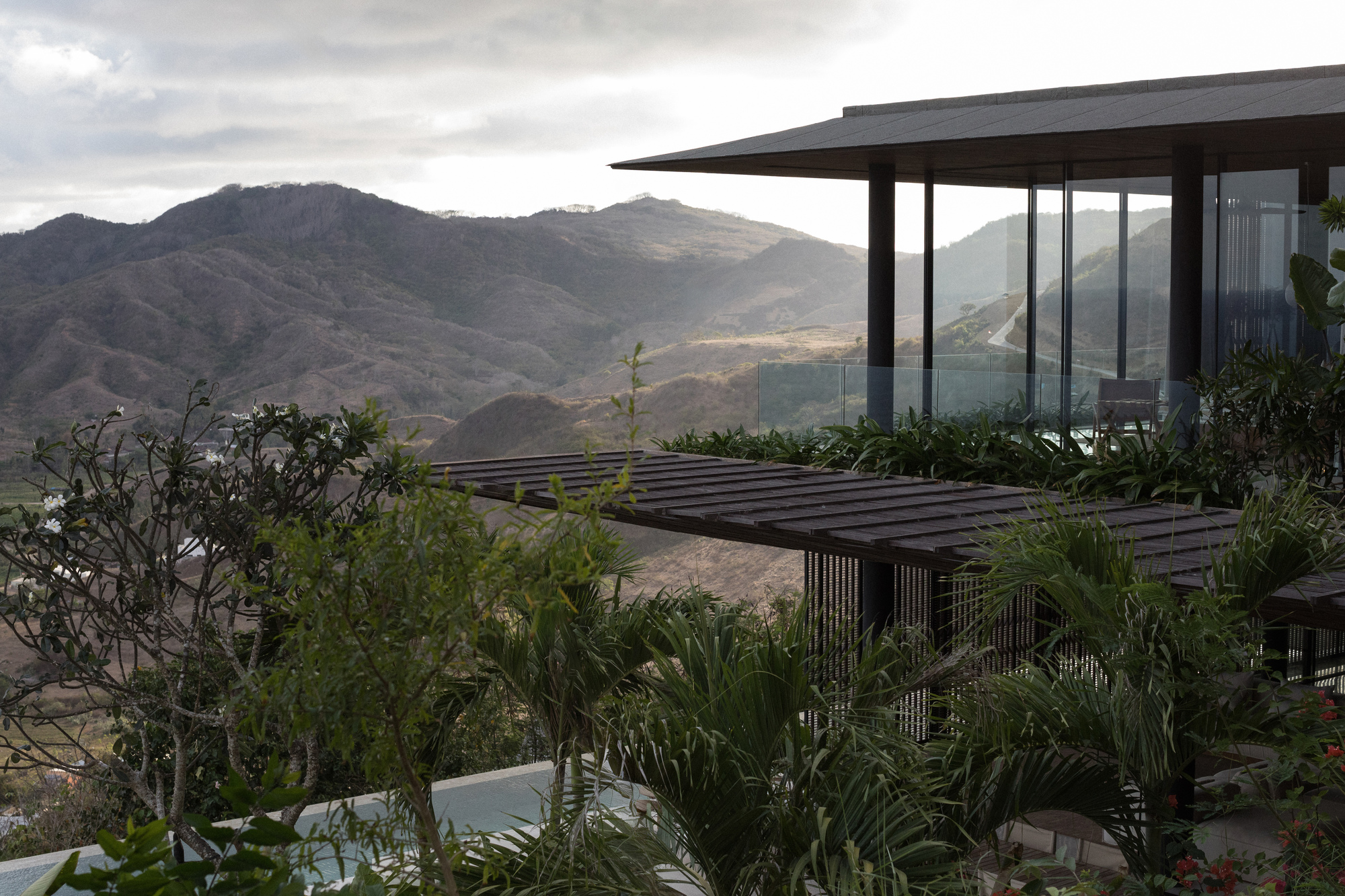Slatted timber pergola and glazed corner pavilion above a pool, framed by palms and frangipani with mountains in hazy light