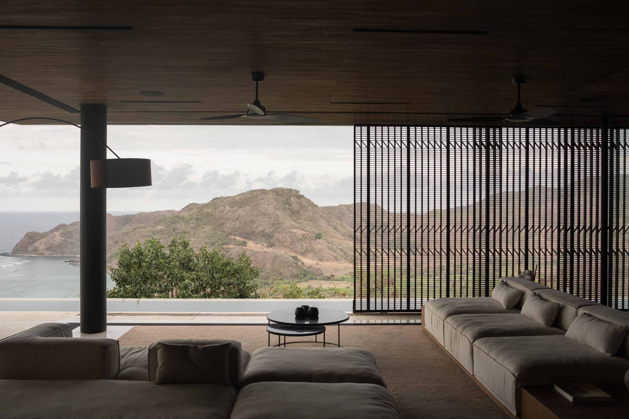 Open living room with low grey sofas, a sliding slatted screen and a steel column framing a bay and hills