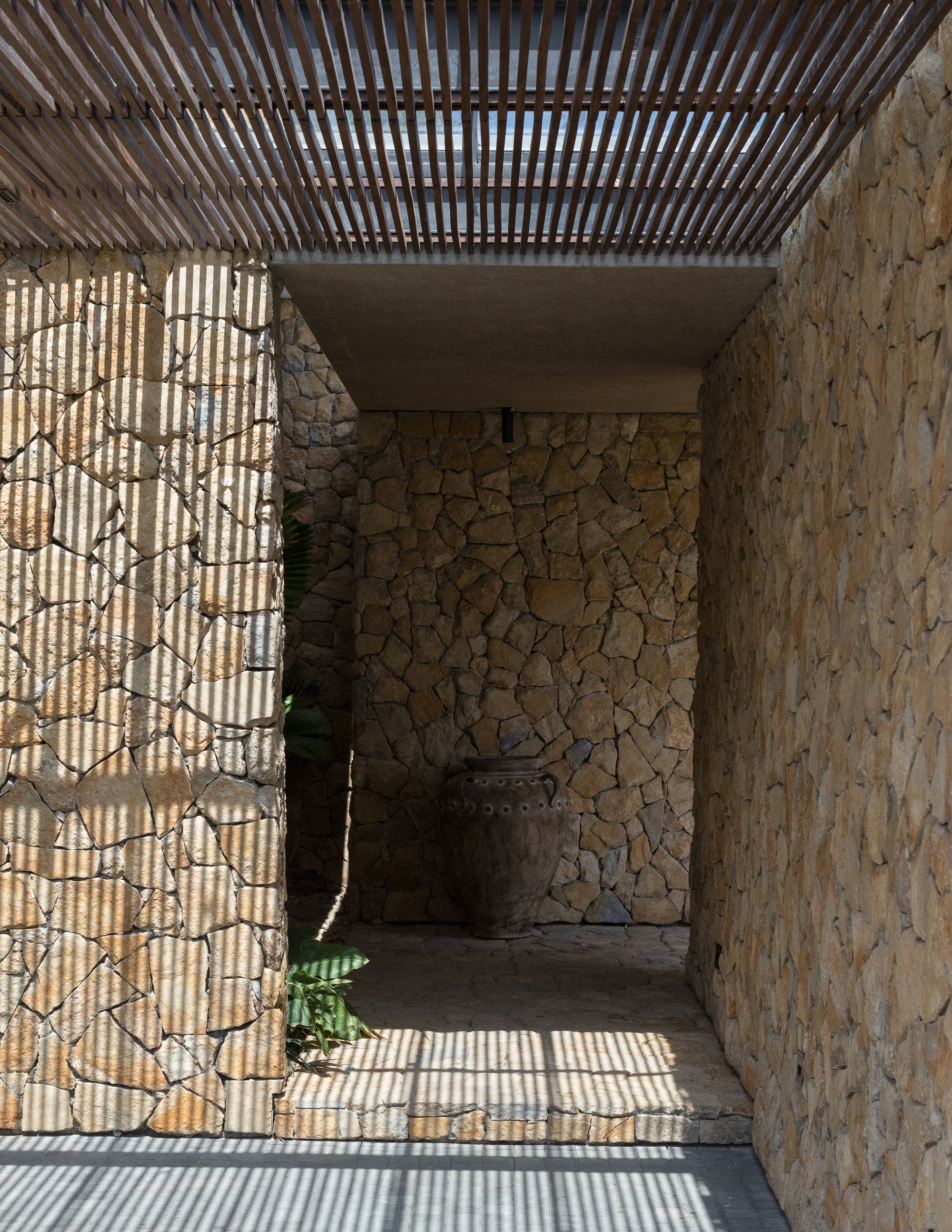 Stone-walled entry passage with a clay urn at the end, striped by sunlight falling through the timber slat ceiling
