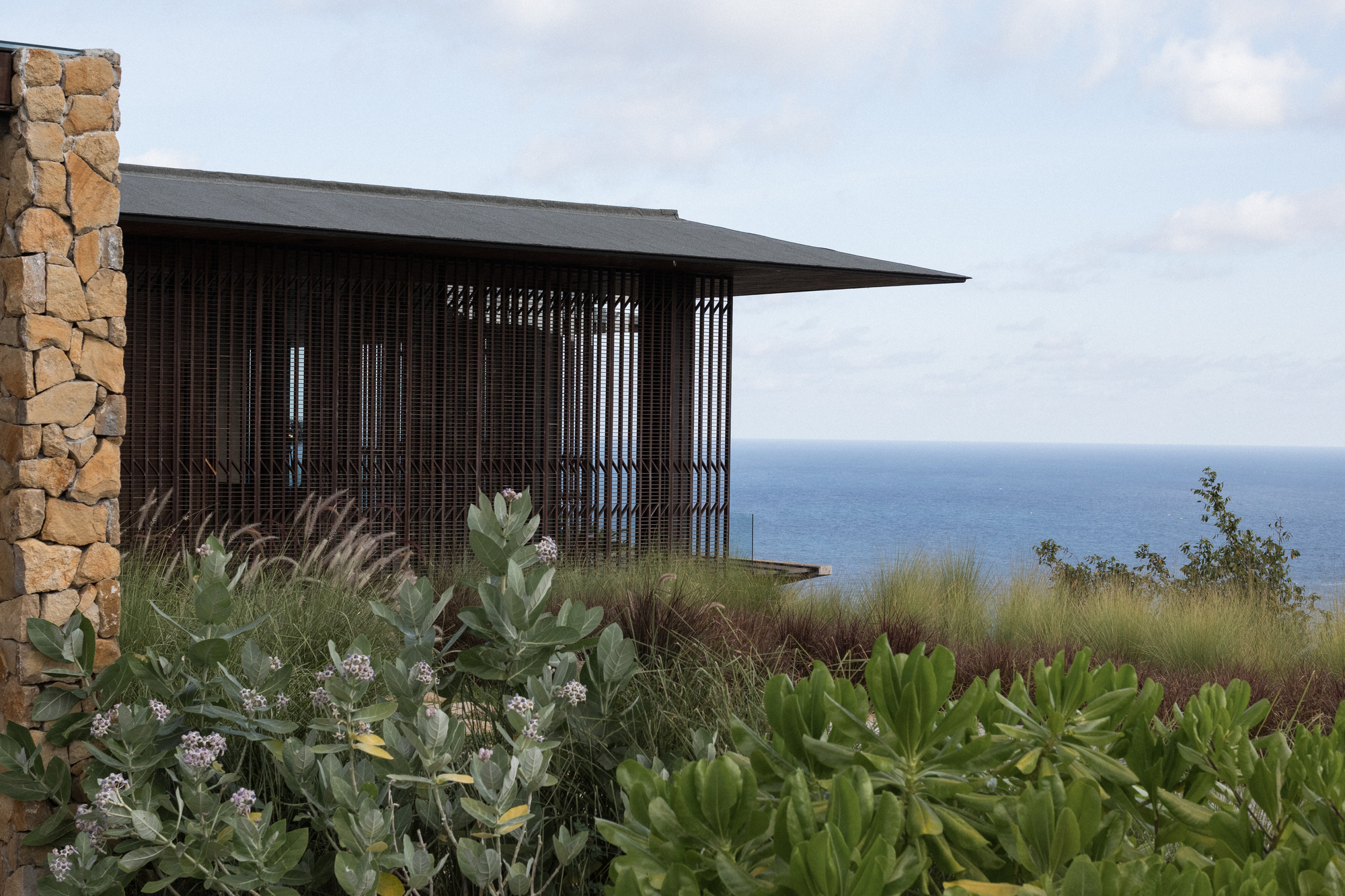 Slatted timber facade under a thin dark roof behind flowering shrubs and grasses, with a stone wall edge and the sea beyond