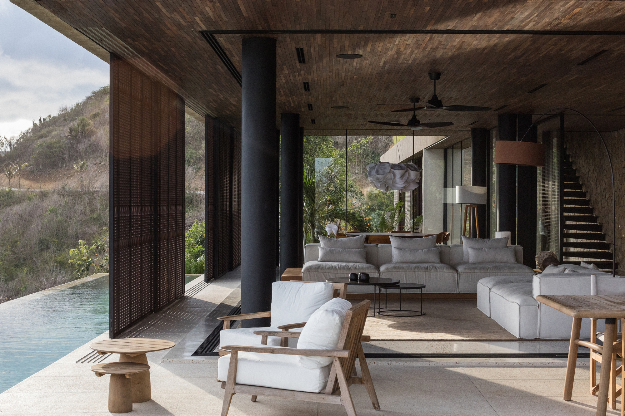 Open living area with timber ceiling, black steel columns, white sofas and teak armchairs beside an infinity pool under a cloudy sky