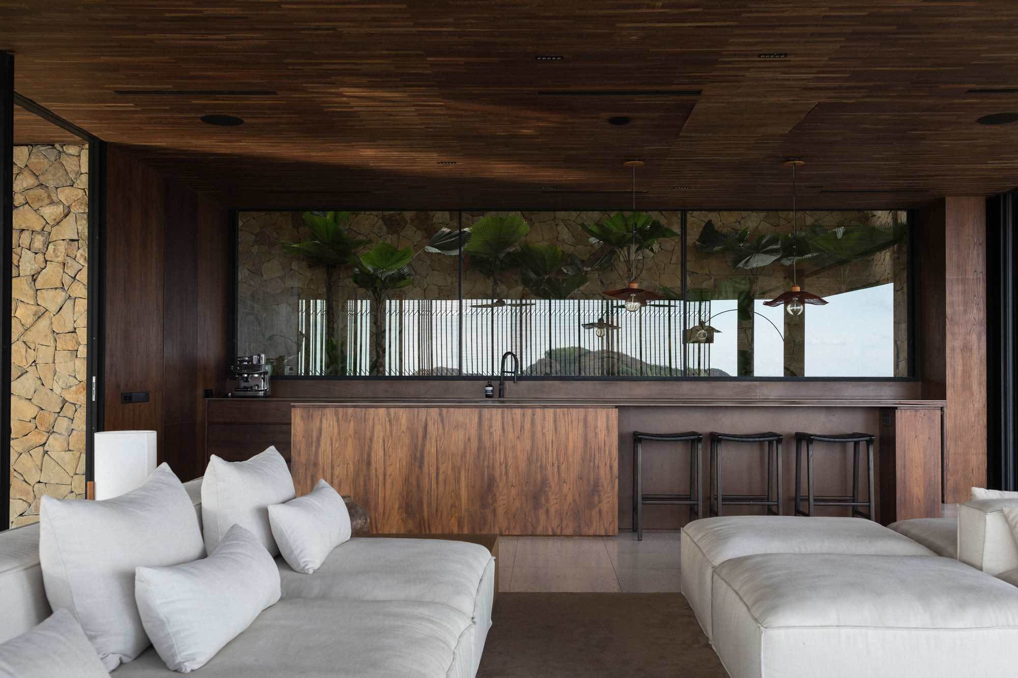 Walnut-clad bar with black stools beneath a slatted timber ceiling, facing a window onto palms and a stone wall