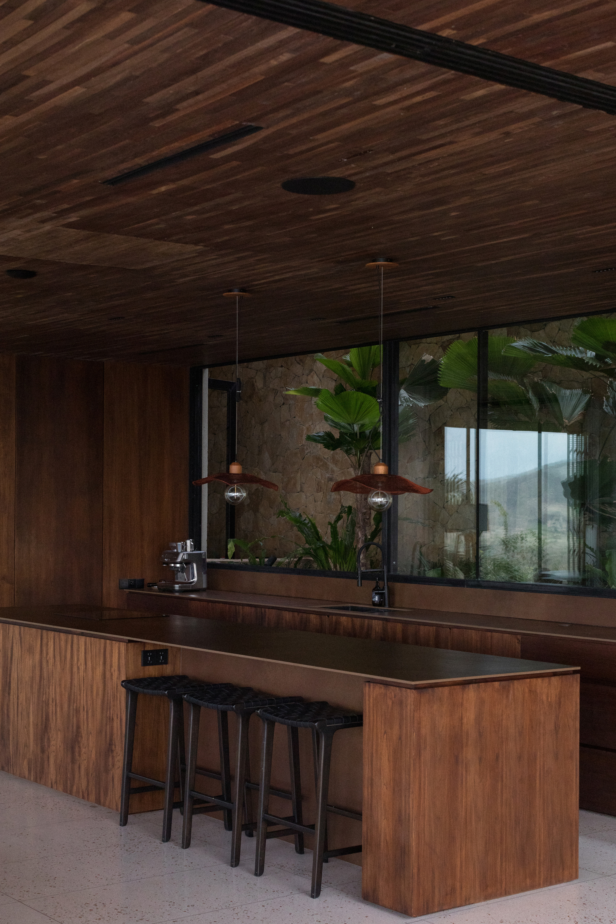 Timber kitchen island with woven black stools and copper pendant lamps, a window of tropical leaves behind in soft daylight