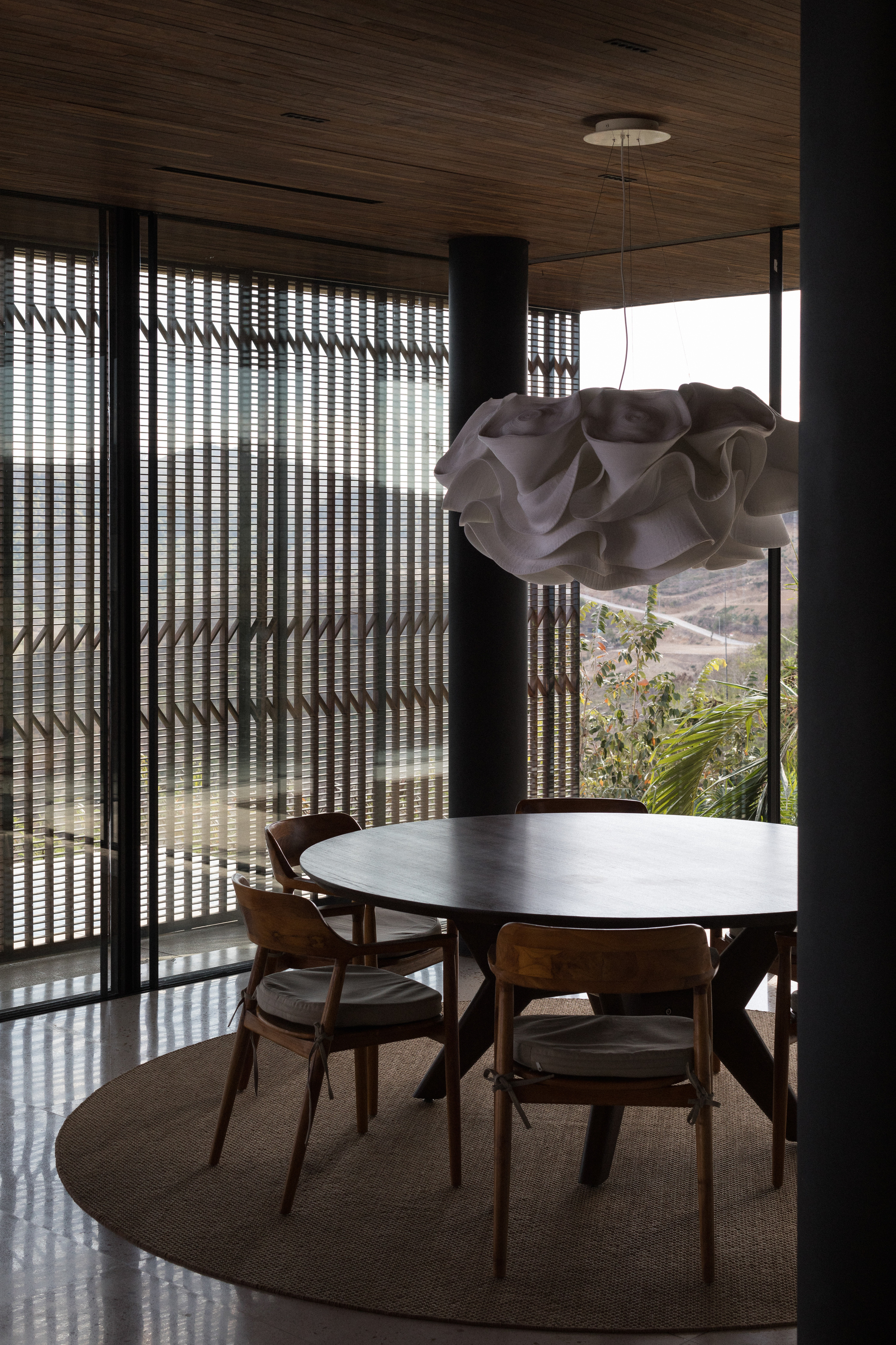 Round dark dining table on a jute rug beneath a ruffled white pendant, with slatted timber screens filtering hillside light