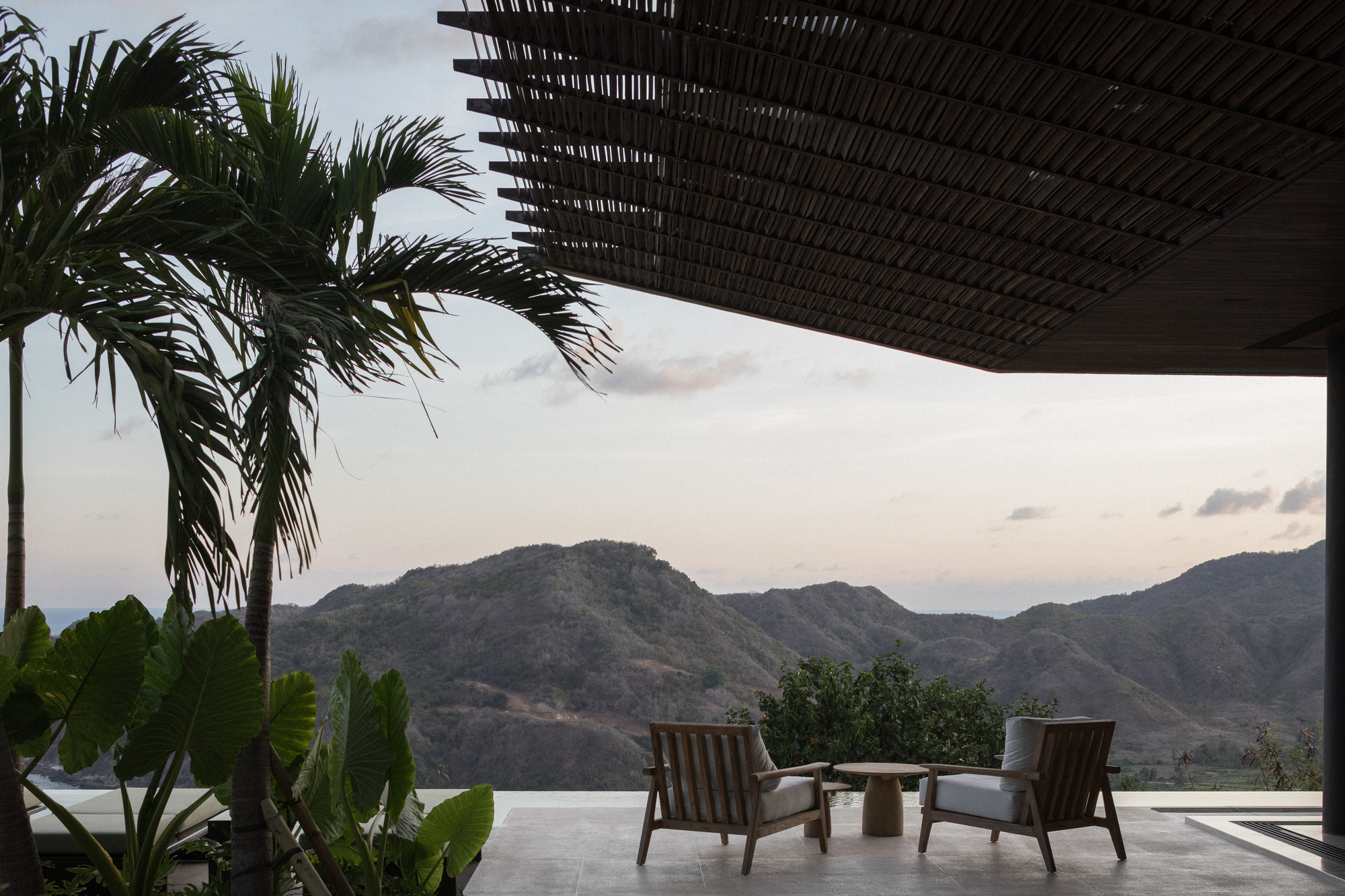 Terrace with two timber armchairs under a slatted canopy, palms in the foreground and dusk light over distant hills