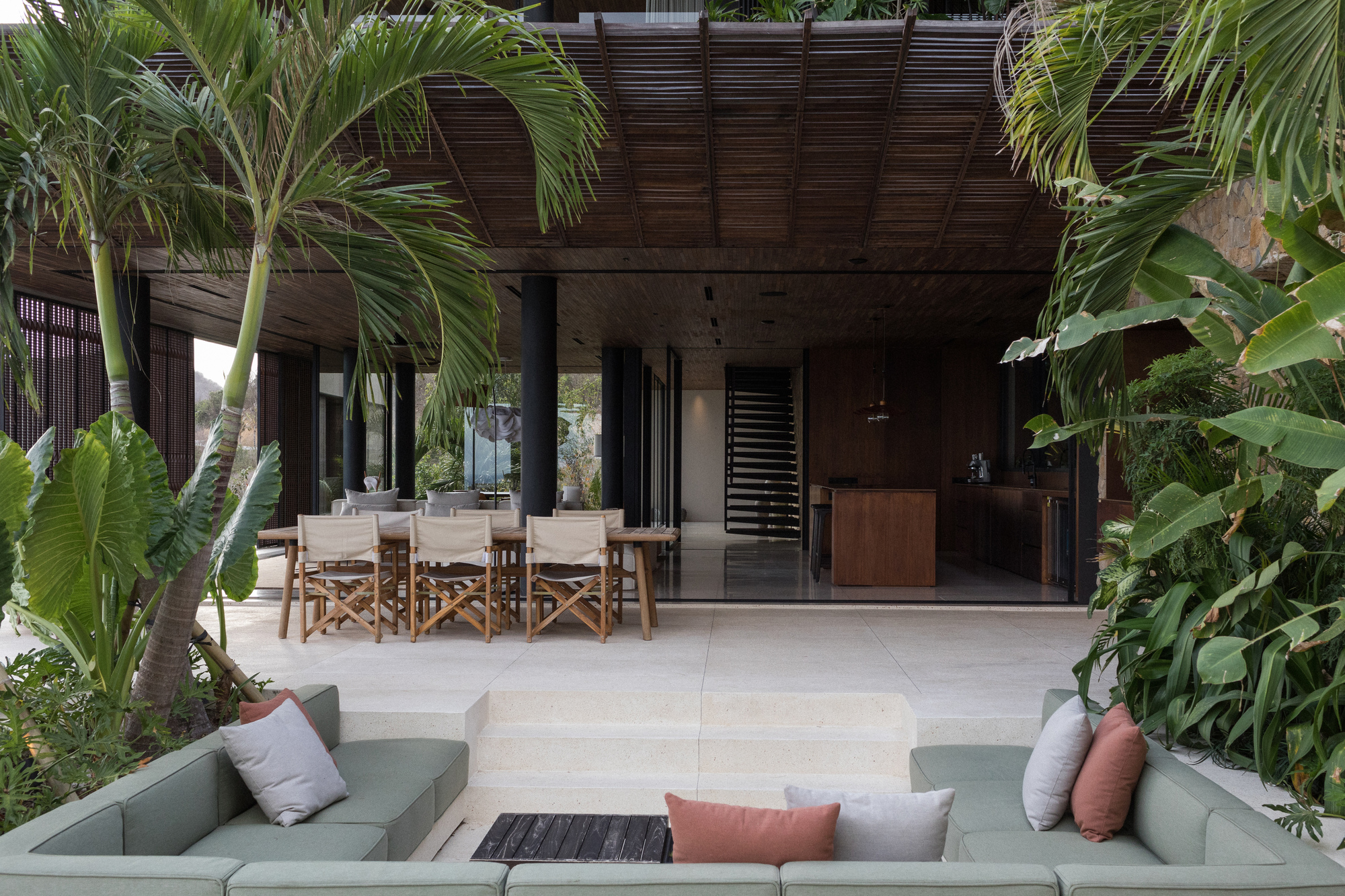 Sunken sage-green lounge in front of an open dining pavilion with slatted timber ceiling, framed by palms and tropical foliage