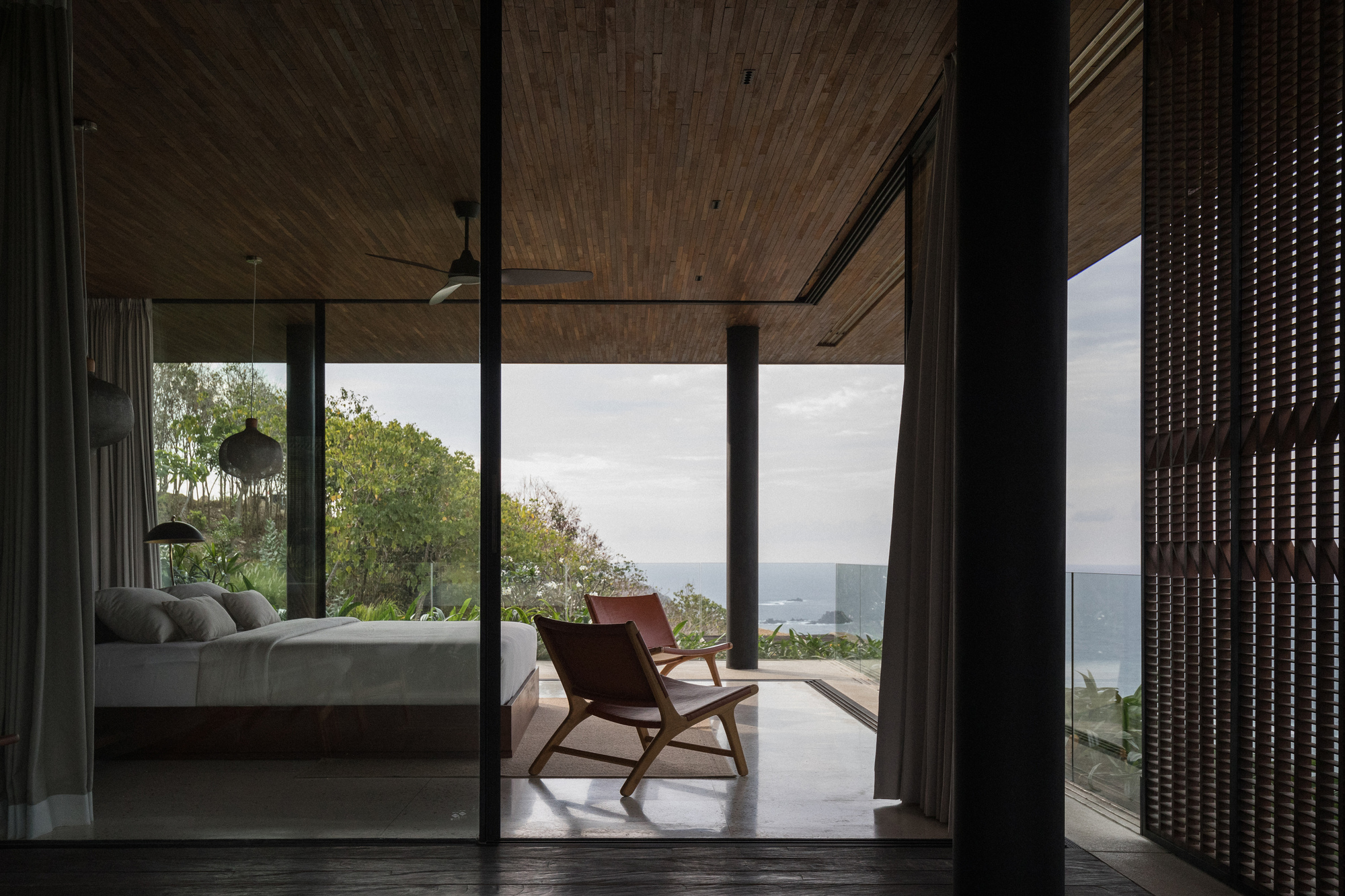 Bedroom with leather chairs and a slatted timber ceiling opening through glass to a gray sea horizon under overcast sky